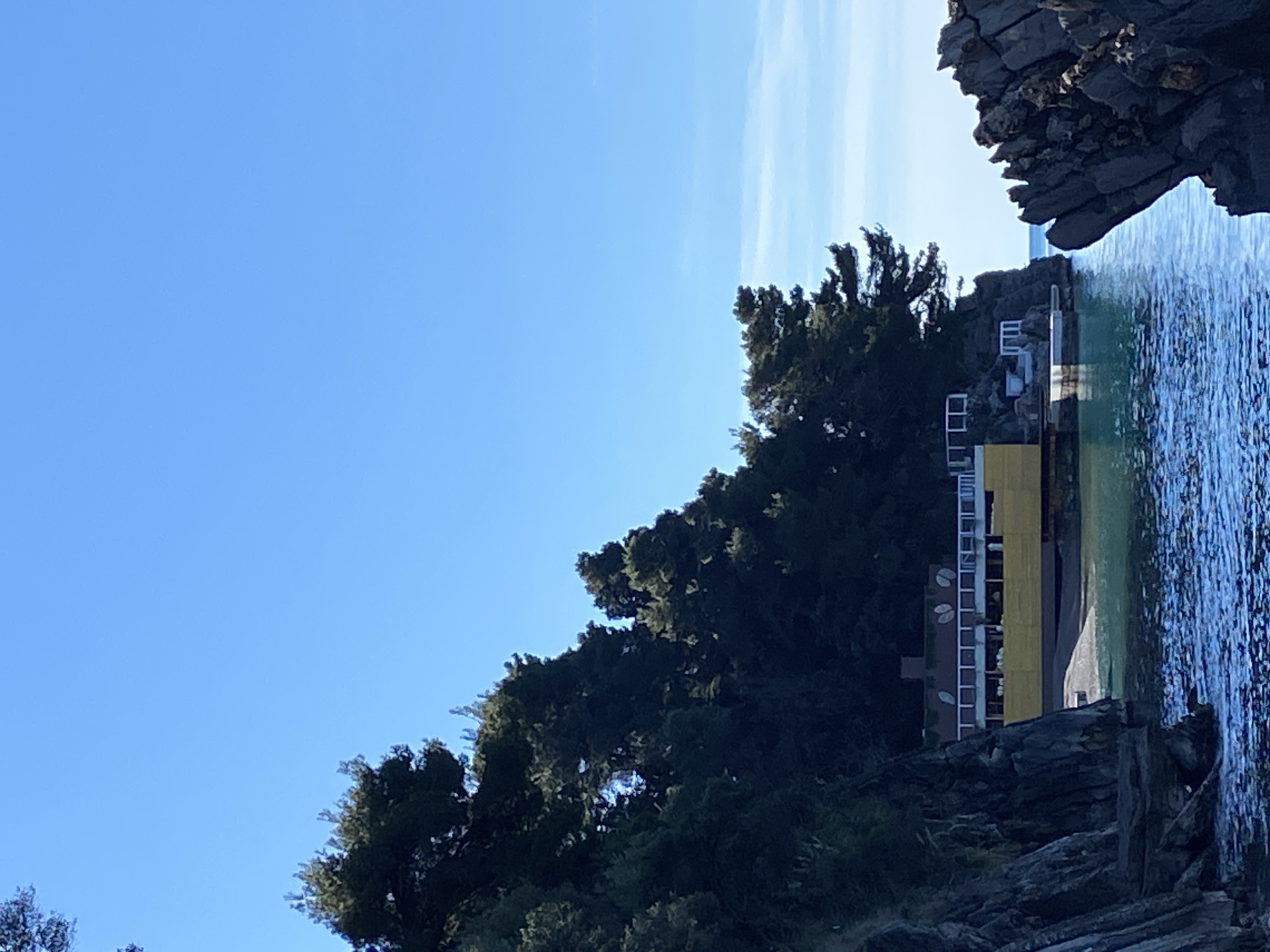 The image depicts a view of a coastline in Budva, Montenegro. The scene includes a cliff formation, a forested area, a building with white railings and yellow walls, and the Adriatic Sea. The sky is clear blue with some faint clouds. The focus is on the coastline where the building is situated. The photograph is taken during daylight hours.