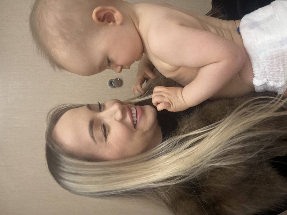 A close-up, rotated image captures a tender moment between a woman and a baby, likely a mother and child. The image is oriented vertically, but the subjects appear to be lying down, interacting horizontally.
If rotated clockwise to an upright position, the woman is on the bottom, lying on her back with her head tilted slightly to her right. She has fair skin, long blonde hair with highlights that spreads out around her head, and is smiling broadly with her teeth showing. Her eyes are gently closed or squinted in an expression of joy and affection.
Positioned above her, and leaning down, is a baby with light blonde hair and fair skin. The baby is shirtless and wears a white diaper. Its small hands are visible near the woman's head and hair. The baby is looking intently down at the woman's face with a gentle, curious, and loving expression.
The background is a plain, light, neutral color, possibly a wall or floor. A small, metallic, circular object, resembling a door fixture or knob, is visible in the background between their heads. The overall scene conveys a warm, intimate, and joyful interaction between the two individuals.