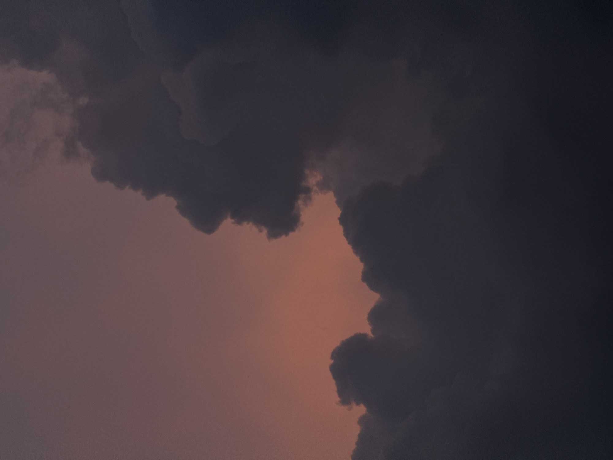 A dusky sky with hues of rose and deep indigo serves as the backdrop for a dramatic formation of dark, heavy clouds. The clouds, dense and textured, occupy the majority of the frame, obscuring much of the sky. Their varied shapes and shadows create a sense of depth and volume. The lighting suggests either early morning or late evening, with a soft, diffused glow emanating from behind the cloud mass on the left, casting a warm, reddish-pink light onto the thinner portions of the clouds and the sky itself. The overall impression is one of a powerful, perhaps impending, weather event.