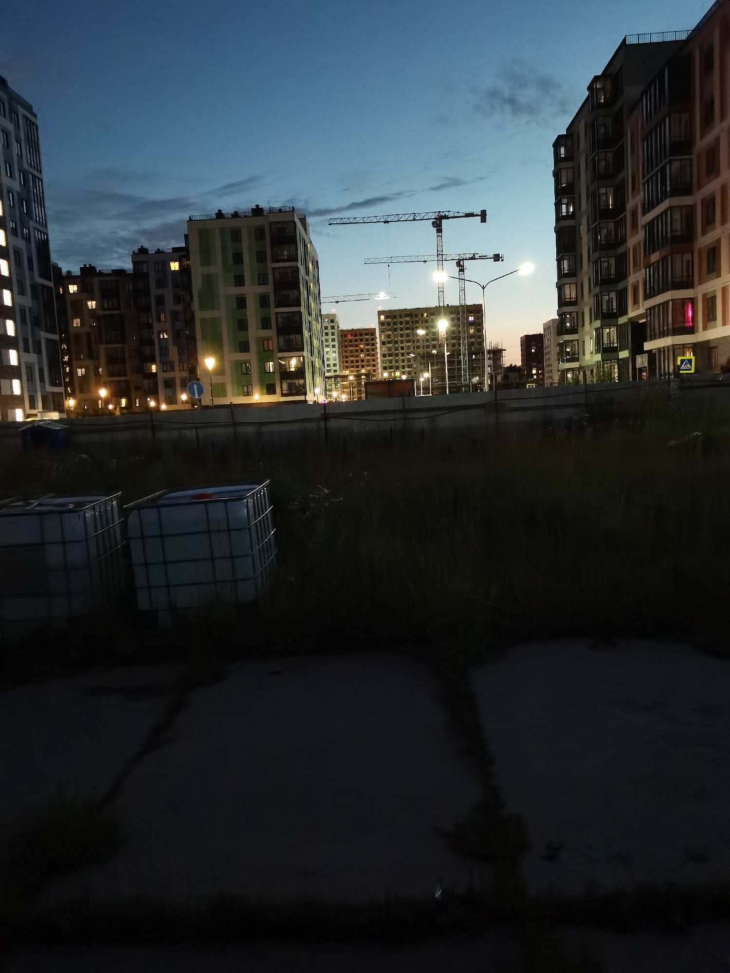 The image captures a nighttime scene in a city, likely a residential or construction area. In the foreground, two large white industrial containers are visible, positioned on what appears to be a patch of dry grass. The background is dominated by several multi-story apartment buildings, some of which are illuminated with interior lights, suggesting occupancy or ongoing work. Construction cranes are prominently featured among the buildings, indicating active development. The sky is dark blue, with hints of clouds and the faint glow of streetlights. A pedestrian crossing sign is visible near one of the buildings, hinting at the presence of public infrastructure. The overall atmosphere is one of twilight or early evening in an urban environment.