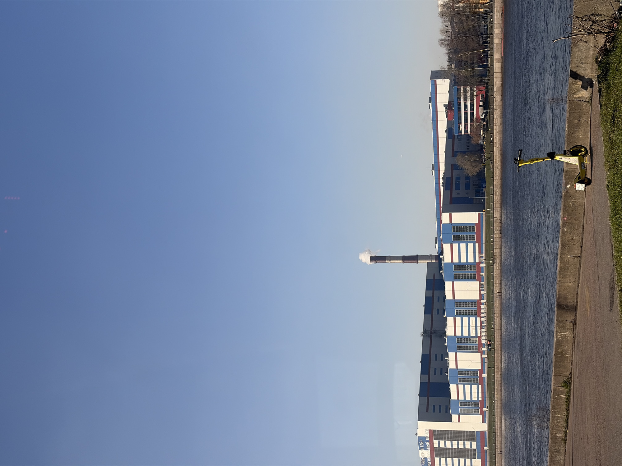 This image, correctly oriented, depicts an urban and industrial riverside scene under a clear blue sky. A calm body of water, likely a canal or river, runs horizontally across the mid-ground.
On the near bank (right side of the image), there is a paved pathway, alongside which green grass grows. A yellow electric scooter is parked on this path, facing the water. The scooter has a license plate or identification tag that is partially visible.
Across the water, on the far bank (left side of the image), stands a large industrial or commercial building complex. The main building is predominantly white and blue, with some red accents, and features multiple rows of rectangular windows. From behind this structure, a tall, slender smokestack rises, emitting a plume of white smoke or steam into the sky. Further in the background, more buildings and bare trees are visible, suggesting a colder season or dormant vegetation.
The clear, bright blue sky indicates good weather and daytime, likely morning or afternoon. No people are explicitly visible in the frame.
Visible text on the side of the main building across the water is in Cyrillic, reading "УНИВЕРСАЛ-НТЕЙ" (UNIVERSAL-NTEY) and "РЕГИОНАЛЬНЫЙ ЦЕНТР" (REGIONAL CENTER). The presence of the smokestack suggests an operational industrial facility or power plant.