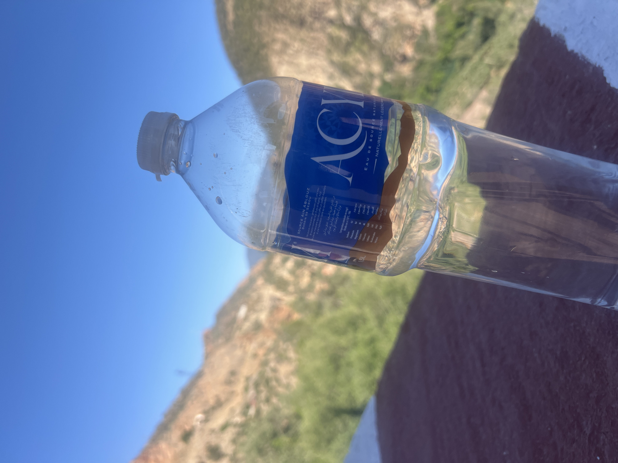 The image depicts a bottle of "ACI Eau de Source Naturellement Légère" mineral water, likely in the Bin El Ouidane, Morocco area. The bottle is partially filled with water, and the label is blue with white lettering. The background shows a hillside with vegetation under a bright blue sky. The bottle rests on a surface with a white edge and reddish-brown coloration. The lighting is bright and sunny, suggesting an outdoor environment.