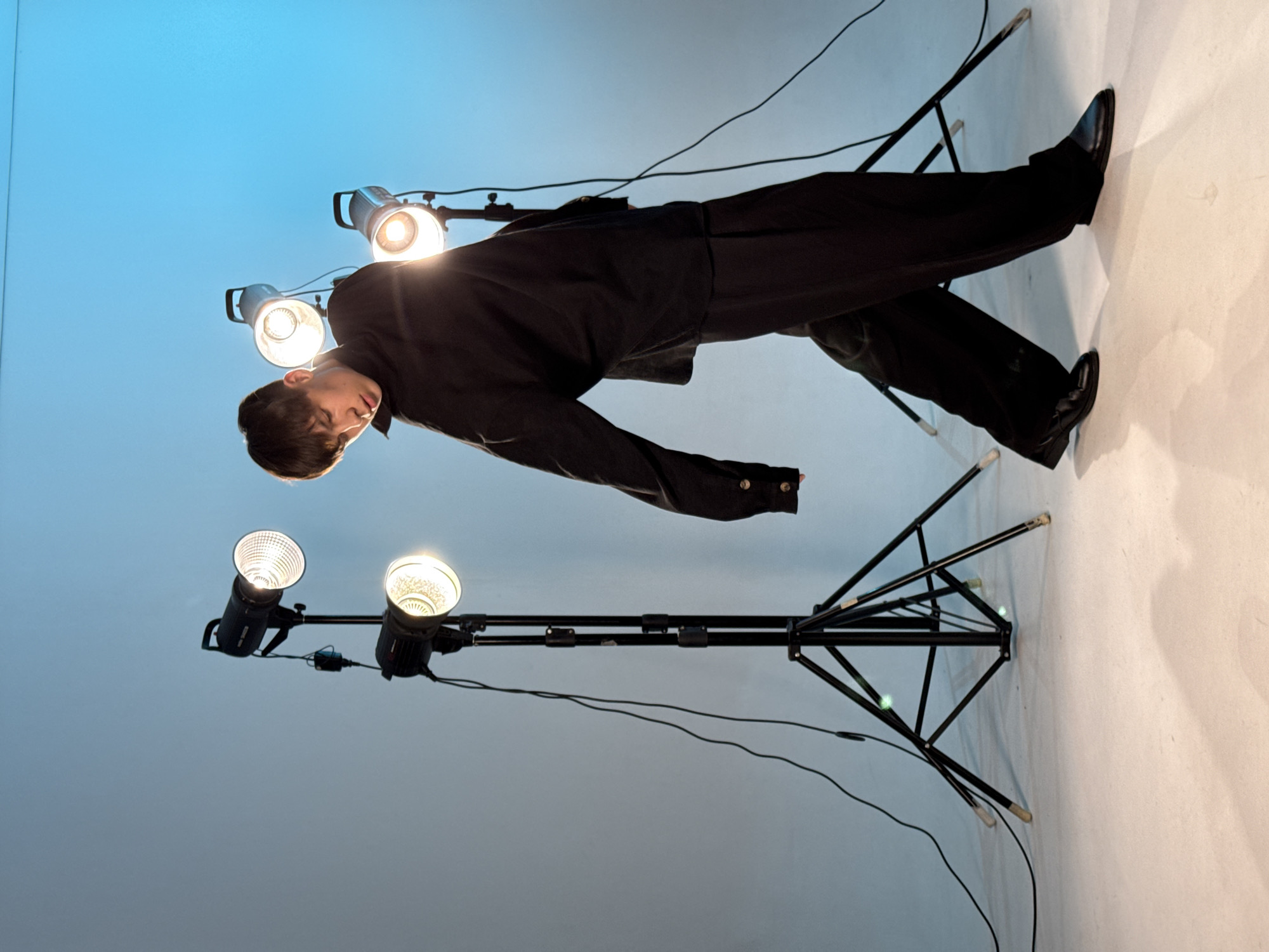 The image shows a man in a dark suit and shoes lying horizontally, seemingly suspended, among several photographic lights on stands. The background is a gradient from blue to white. The lights are mounted on black stands, and power cords are visible. The location in the city of Chaikovsky, Russia, is not discernible from the image. The details are focused on a studio setting with lighting equipment and a man in formal attire.