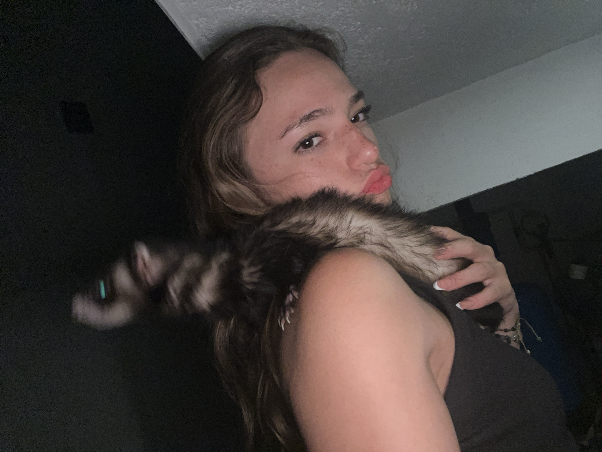 A young woman is pictured indoors, holding a ferret around her neck. The woman has brown hair, freckles, and is pouting her lips. She is wearing a black tank top. The ferret, with dark brown and black fur and a bushy tail, is positioned on her shoulder. The background is dark, with a glimpse of a white ceiling and wall. There is nothing in the image to indicate it was taken in San Bernardino Tlaxcalancingo, Mexico.
