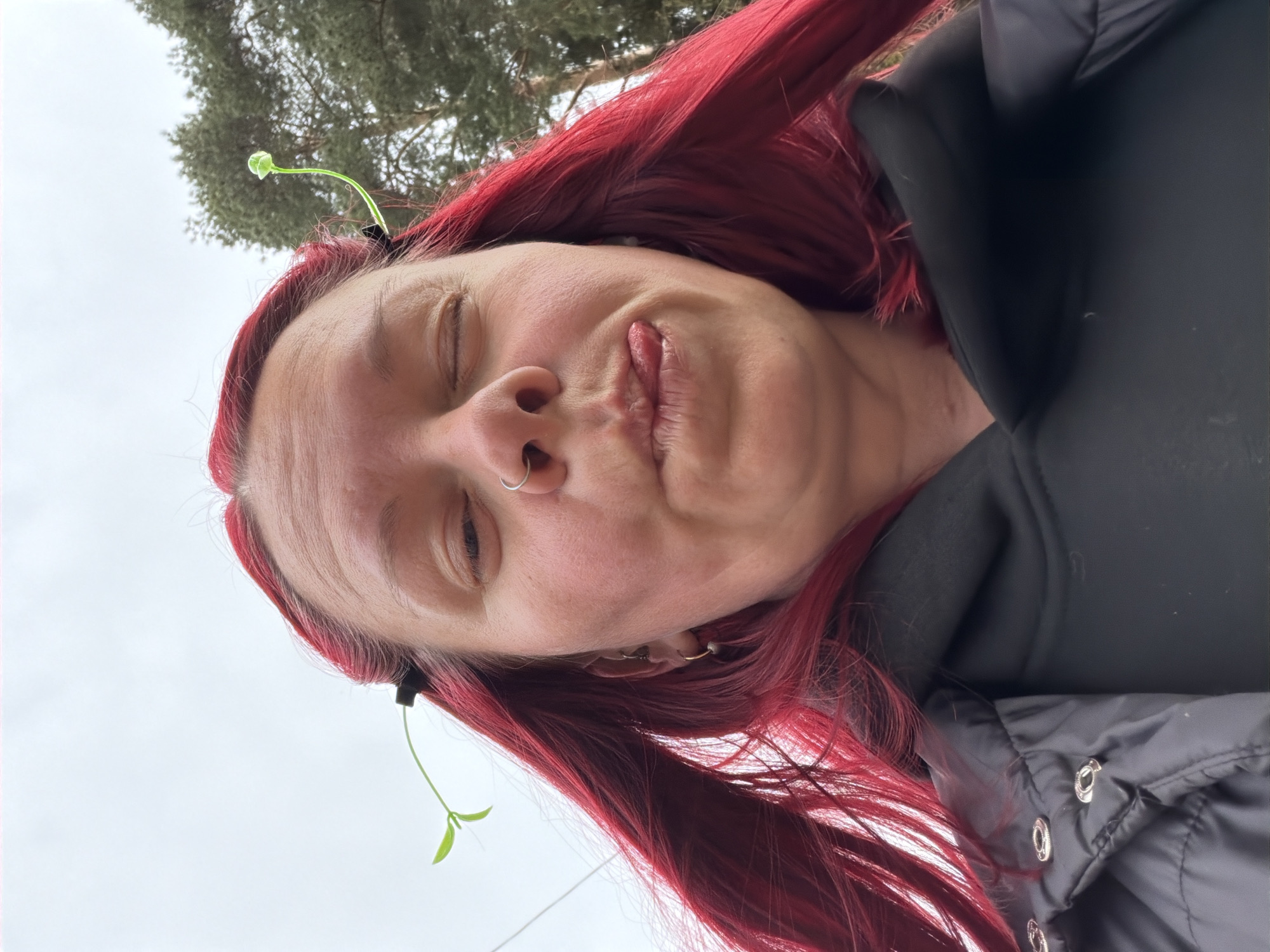 The image is a close-up selfie of a fair-skinned person with long, dyed red hair. A nose ring is visible. There are two green sprouts placed in the person's hair near the temples. The person is wearing a dark-colored jacket. The background shows a cloudy sky and a tree. Based on the information provided, it is assumed that the location is Chaikovsky, Russia.