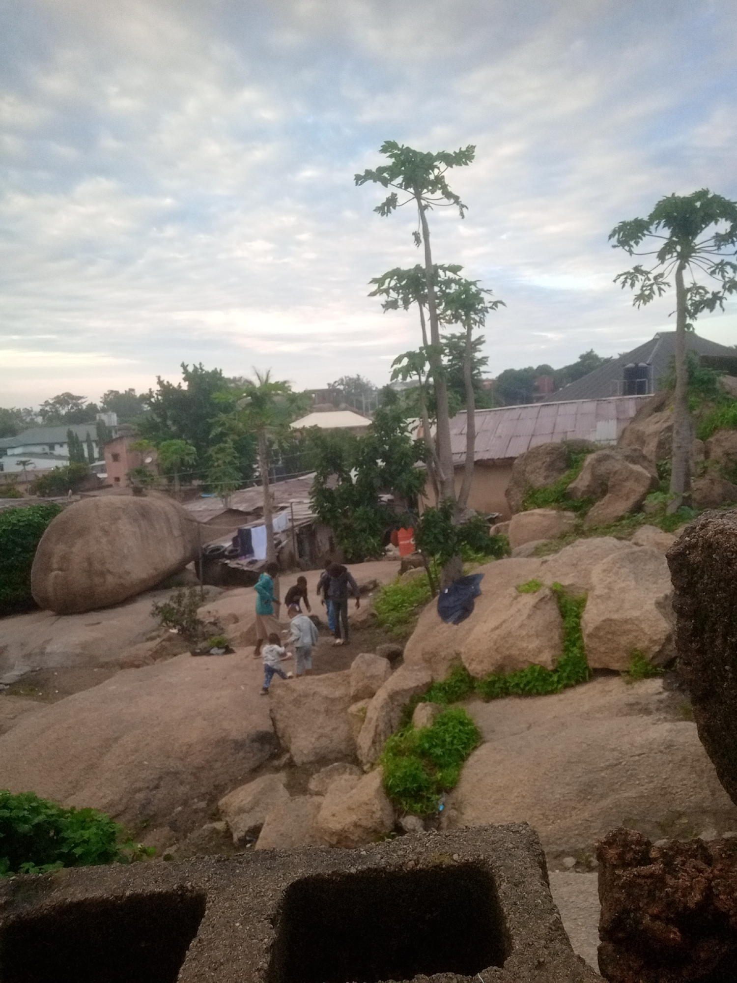 An outdoor, high-angle, medium shot captures a rocky, uneven terrain in what appears to be a residential area with sparse vegetation. The sky is overcast with soft, diffused light. In the foreground, the top edges of two concrete structures with a rough texture are visible. The midground is dominated by large, tan-colored boulders scattered across the landscape. A group of at least five individuals are present. Two adults, one wearing a teal shirt and the other a dark jacket, are walking uphill towards the right. Two children in lighter clothing are also walking uphill to the right, one slightly ahead of the other. Another child, wearing a blue shirt and jeans, is standing and looking towards the right. A dark blue plastic bag rests on a boulder to the left of the group. In the background, several buildings with corrugated metal roofs are visible, along with scattered trees and foliage. A large, rounded boulder, roughly the size of a small vehicle, is situated to the left of the group of people. Clothes are hanging on a line between two structures on the far left. A few tall, slender trees with sparse foliage, resembling papaya trees, are scattered throughout the scene.