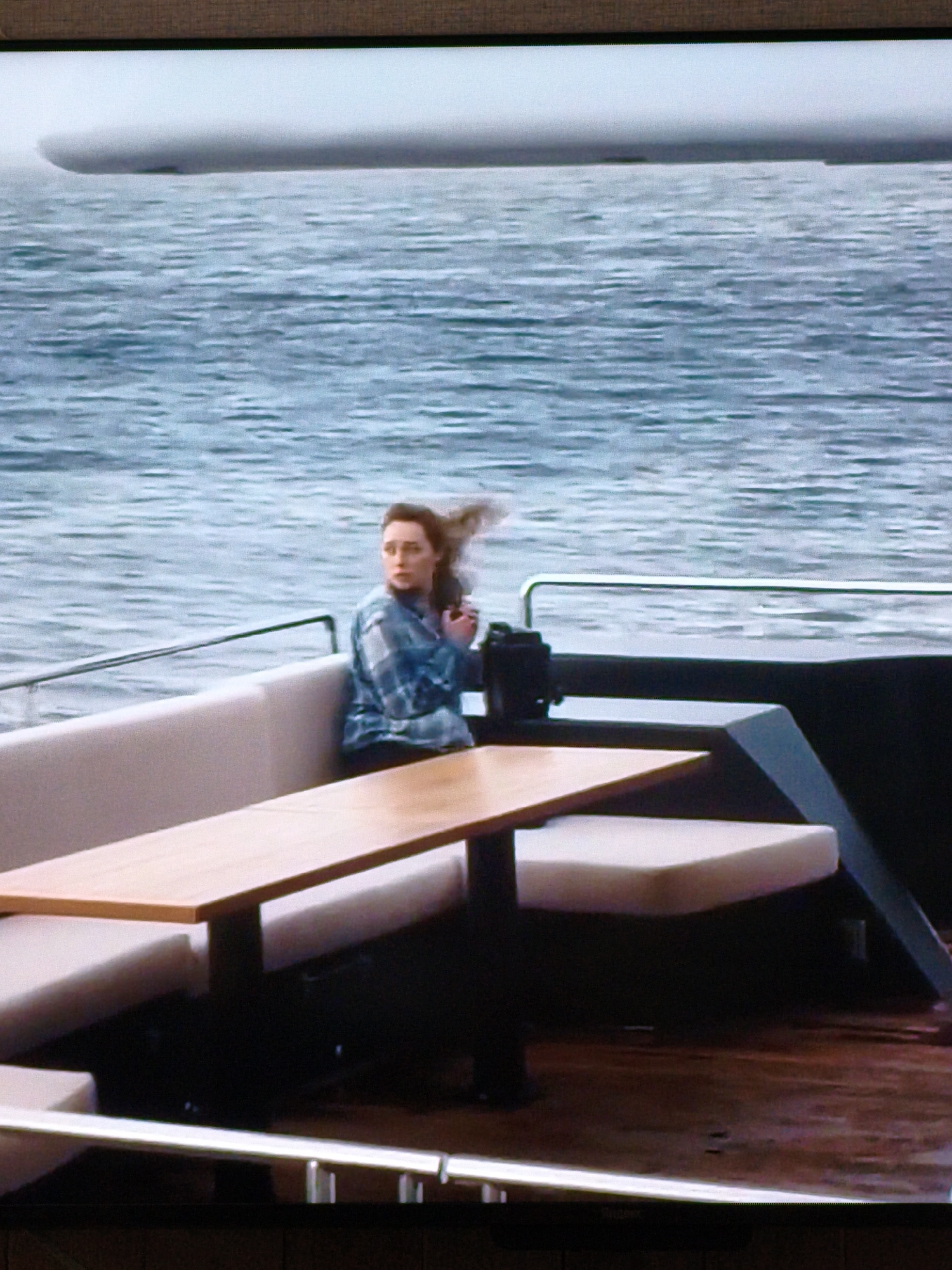 A woman is sitting on a boat with a table in front of her. The boat is on the water, and there is a large object in the background. The woman is wearing a blue and white plaid shirt and has her hair pulled back. She appears to be looking to her right. The table is made of wood and has a light-colored surface. The boat has white seating and a dark railing. The water is choppy, and the sky is overcast. There are no recognizable landmarks that indicate the location in Vladivostok, Russia.