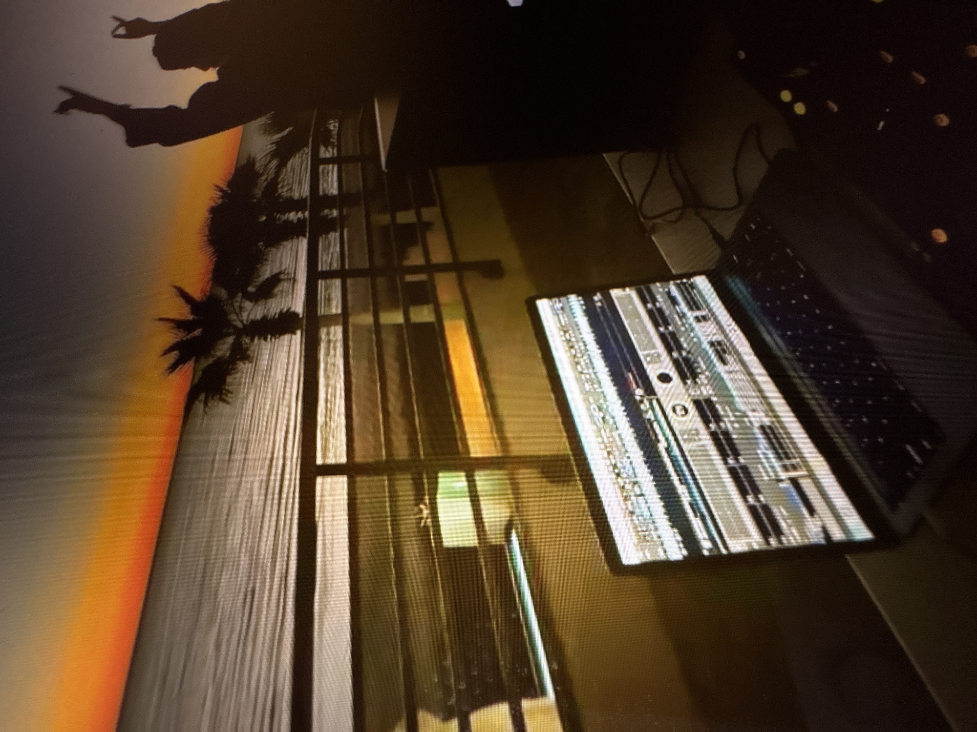 The image depicts a silhouetted scene of what appears to be a DJ setup at sunset, with the ocean and palm trees in the background. A laptop is open on a table, displaying a waveform or audio mixing software. To the right, a large black speaker is visible. In the background, two silhouetted figures are raising their hands, one in a gesture resembling the "rock on" sign. The sunset casts an orange and grey glow over the water and the sky, with palm trees silhouetted against the horizon. The railing of a boardwalk or pier is in the foreground, separating the setup from the water. The scene is likely set at a beach or waterfront location. There is no information available to determine the location in Pori, Finland.