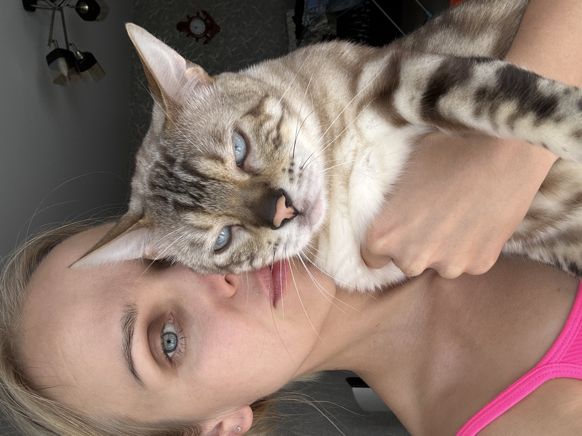 A close-up, selfie-style image features a young Caucasian woman with blonde hair and blue eyes, holding a grey and white tabby cat. The woman is wearing a bright pink tank top and is looking directly at the camera. Her left arm is wrapped around the cat, which is held close to her face. The cat also has blue eyes and is looking slightly to the left of the camera. The background is dimly lit and appears to be an indoor setting, possibly a living room, with a patterned wallpaper and a light fixture visible on the ceiling. There is no indication of the specific location within the city of Energetik, Russia.