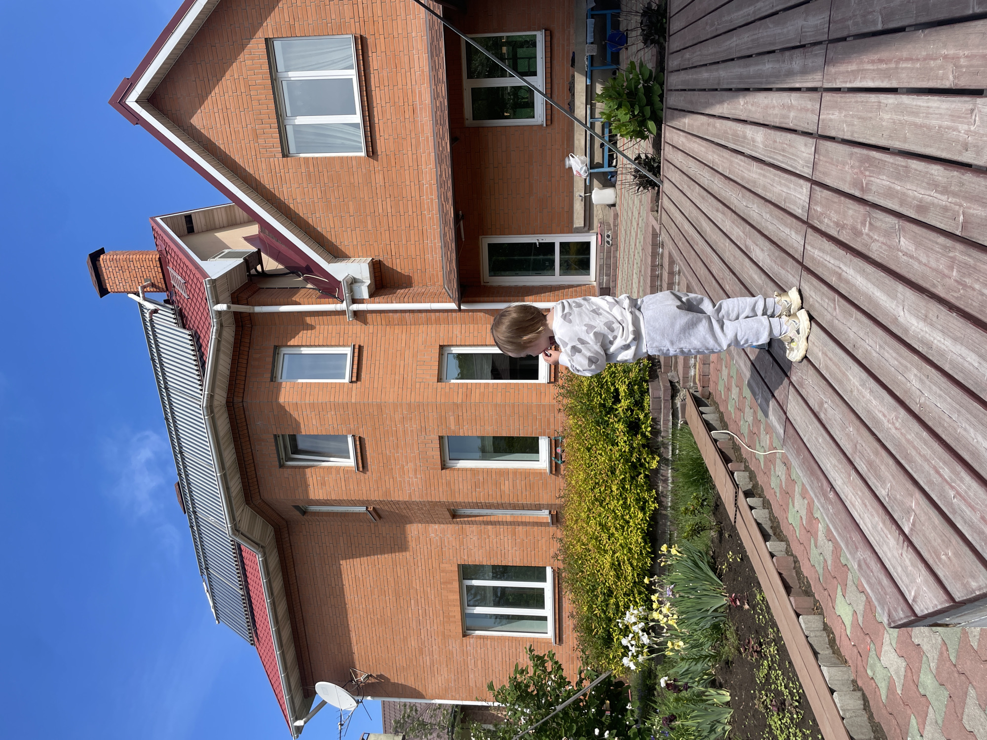 A young child in a white sweat suit stands on a wooden deck adjacent to a two-story red brick building under a clear blue sky. A flower bed runs along the edge of the deck near the base of the building. A satellite dish is mounted on the side of the building. The general location is Vladivostok, Russia.