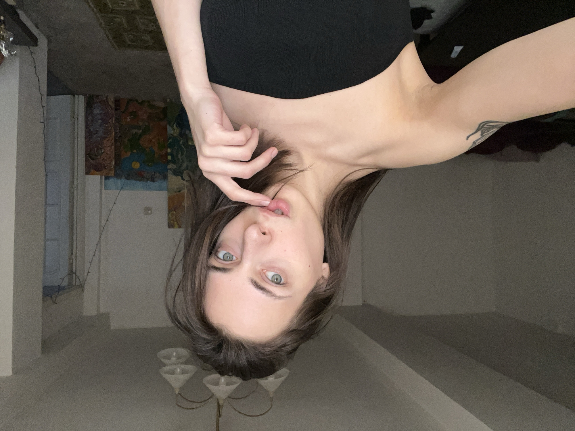 A woman with brown hair and light eyes is positioned upside down in a room. She is wearing a black top. Her left hand is near her face, with a finger touching her lip. A tattoo is visible on her right arm. In the background, there is a chandelier and walls, with an image of a painting on the wall. The location in Krasnoyarsk, Russia is not possible to ascertain.