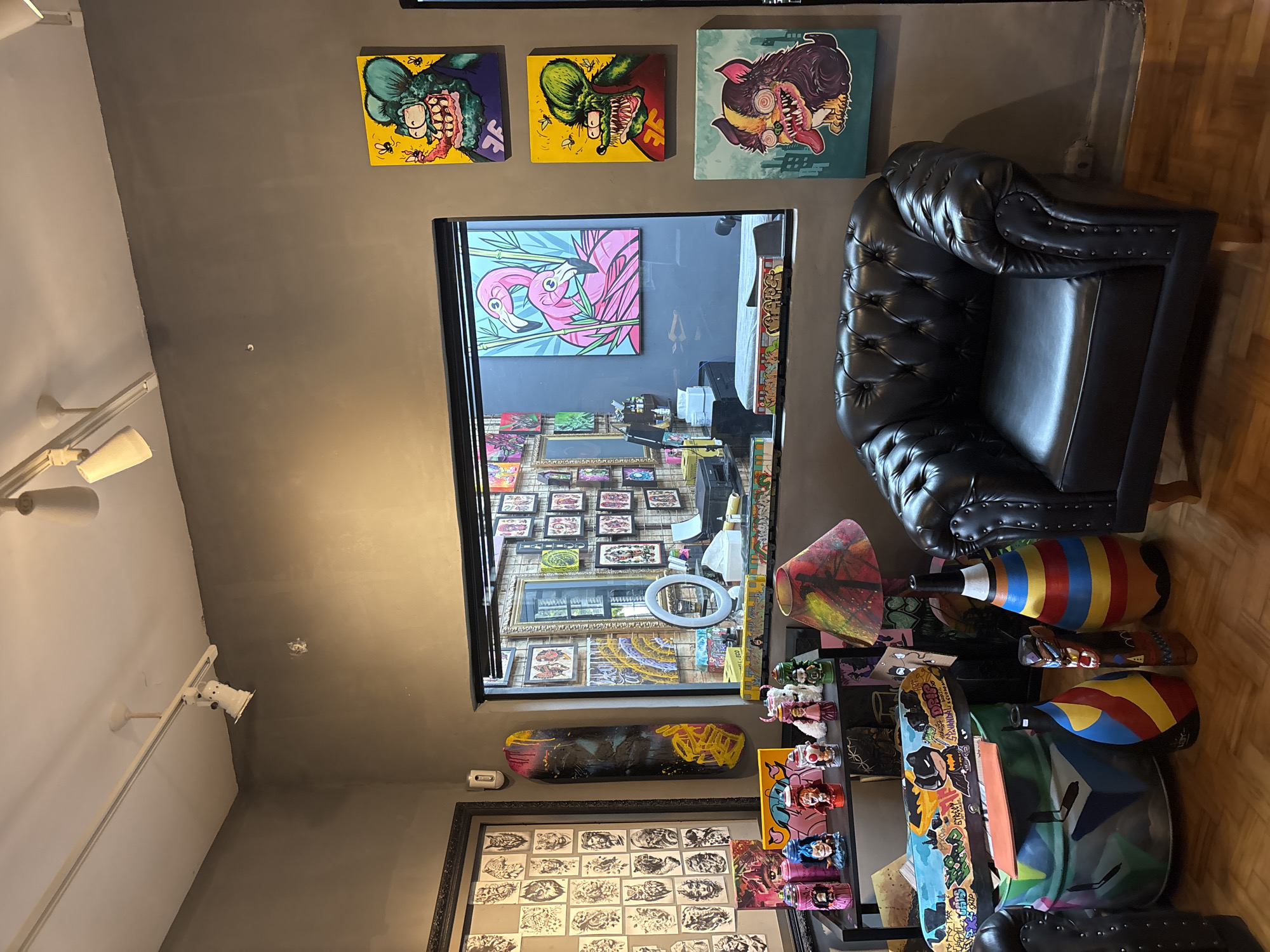 The image shows a room interior, possibly a tattoo parlor, in São Paulo, Brazil. The walls are a neutral gray. Above a large rectangular window that looks into another room are three framed cartoonish images of monsters. The window reveals a room with framed artwork on the walls and a pink flamingo picture visible. To the right of the window is a black leather tufted chair. Below the window is an assortment of skateboards, painted wooden figures, and other decorative items. Below these is a framed collection of black and white tattoo sketches and more artwork and skateboards. Track lighting is visible on the ceiling. The floor is wooden.