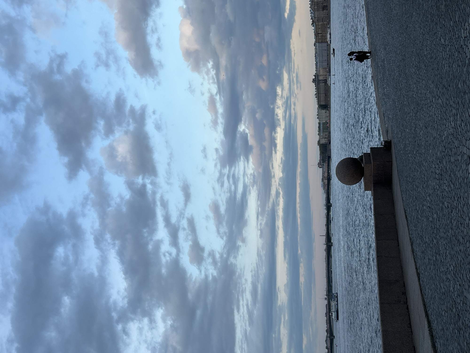 The image depicts a cloudy sky over the Neva River in Saint Petersburg, Russia. A stone embankment with a decorative granite ball runs along the right edge. Two figures are visible further down the embankment. Buildings line the opposite bank of the river. Boats are visible on the river. The embankment is paved with cobblestones.