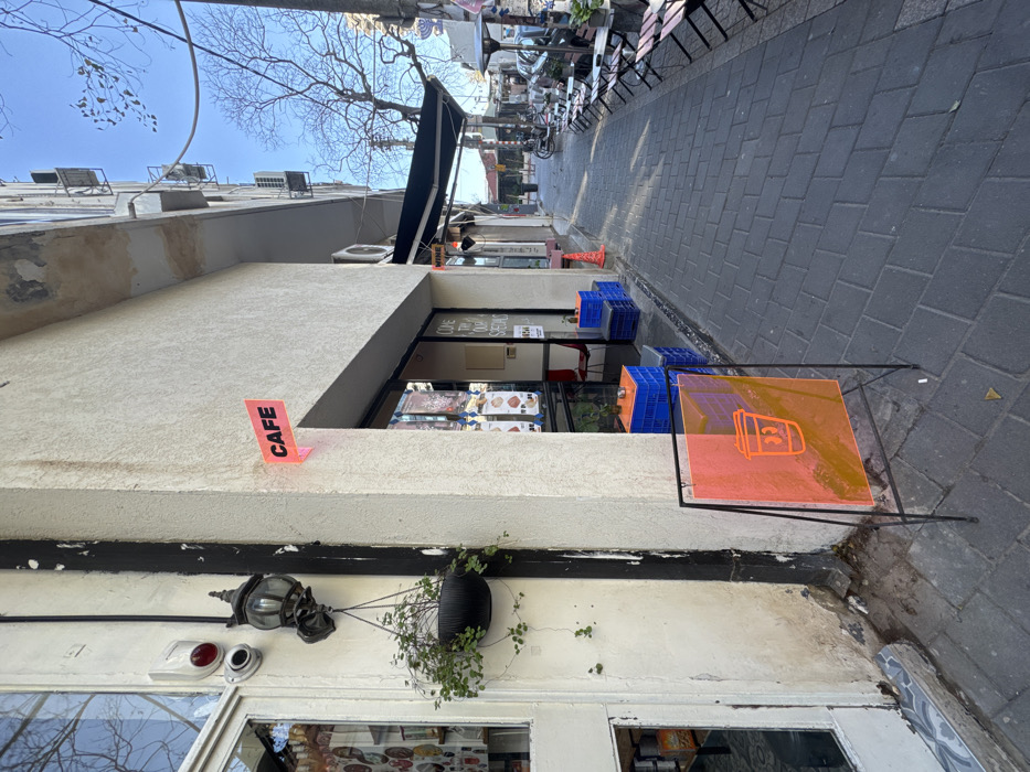 A cafe exterior features a small, street-facing window with visible pastry displays. A pink "CAFE" sign is affixed above the window. Blue crates and an orange sign with a coffee cup graphic sit on the sidewalk. Above the cafe entrance is a hanging plant and decorative lighting. The street narrows with outdoor seating visible in the distance. The sky is pale blue with bare tree branches overhead.