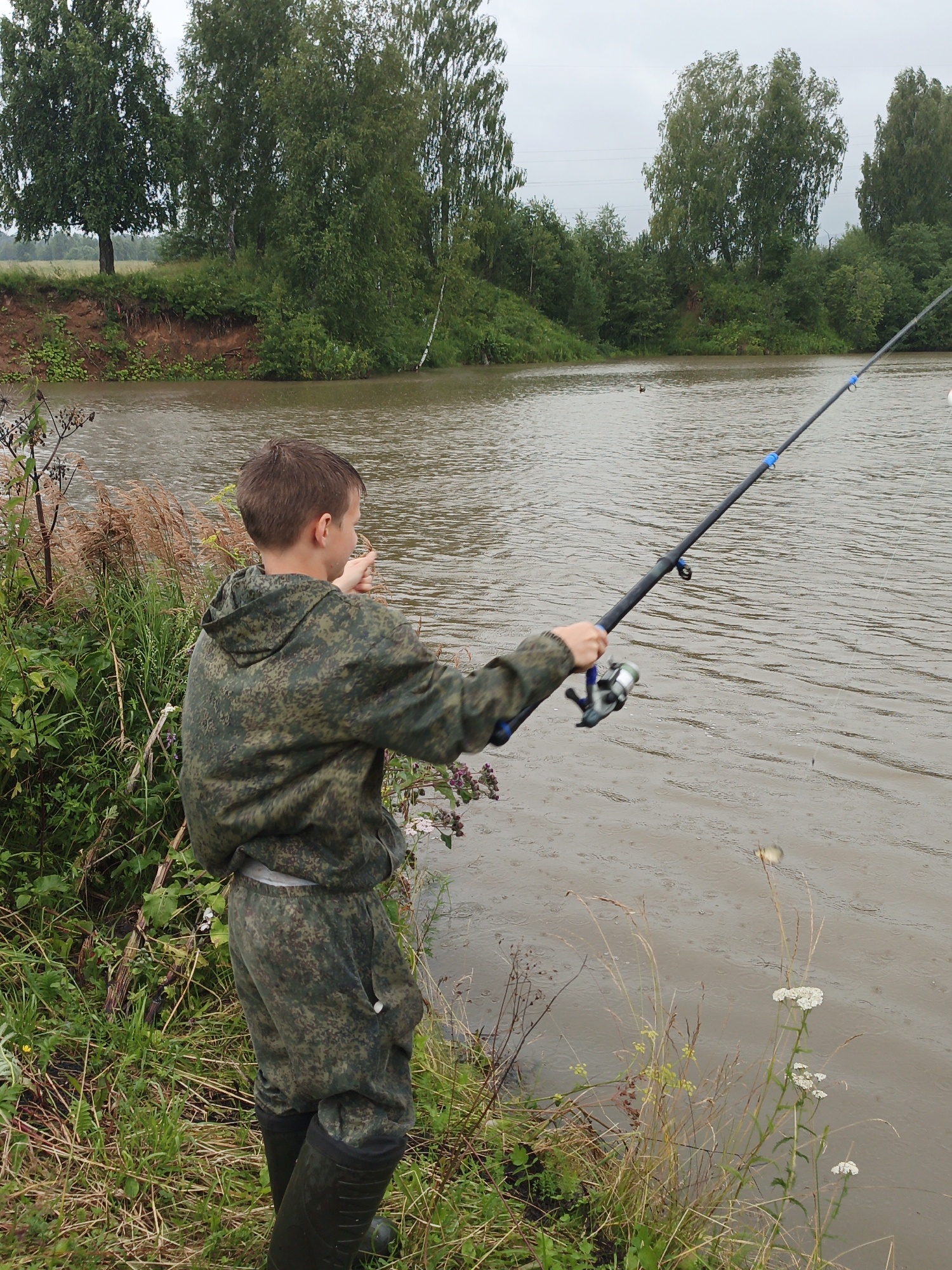 The image shows a young male, facing away from the camera, fishing on the bank of a river in Urtiushka, Russia. He is wearing a camouflage suit and black rubber boots, and he is holding a fishing rod. The river is wide and muddy, with trees and vegetation lining its banks. The sky is overcast, suggesting a cloudy or rainy day. The overall scene depicts a peaceful, outdoor activity.