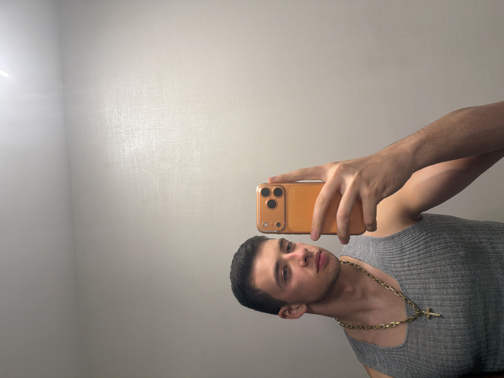 A young man is taking a selfie indoors. He is holding an orange-colored smartphone. He is wearing a grey ribbed tank top and a gold chain with a cross pendant. The background is a plain, light-colored wall. The location is unknown.