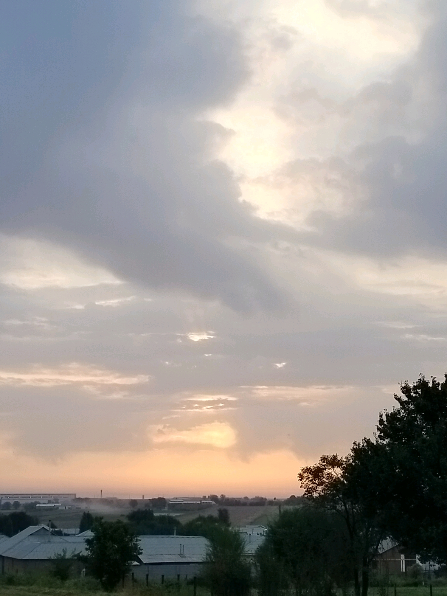 The image depicts a panoramic view of a landscape at dusk, with the sky dominating the upper portion of the frame. Large, diffuse clouds are present, with a bright, diffused light source, likely the setting sun, visible towards the center of the image, casting an orange and pink hue across the lower sky. The foreground reveals a rural or semi-urban setting with several low-rise buildings with metal roofs. Vegetation, including trees and bushes, is scattered throughout the scene. In the middle ground, there appears to be industrial or agricultural structures in the distance, with some haze or mist visible. The overall impression is one of a tranquil evening in a locality that combines residential, industrial, and natural elements. The location is specified as the city of Көлкент, Kazakhstan. There are no people or animals visible in the image.