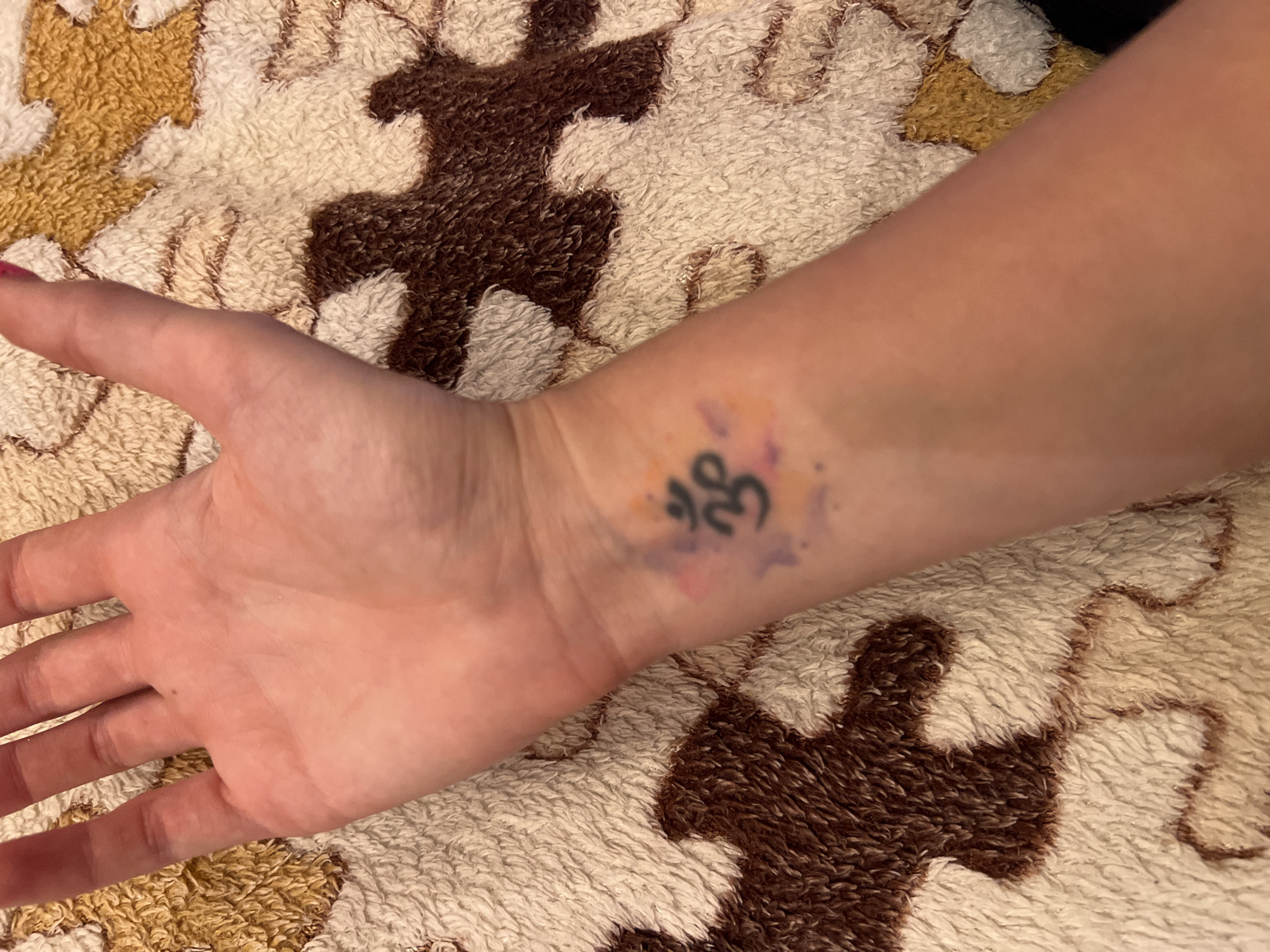 The image shows a close-up view of a person's left forearm and hand, resting on a textured, patterned fabric that appears to be a blanket or towel. The fabric has a fuzzy texture and features a repeating pattern of brown and beige shapes. On the forearm, there is a small tattoo of the Om symbol, rendered in black ink, with subtle watercolor-like splashes of purple and orange surrounding it. The skin of the arm is fair, and the hand, palm facing upwards, shows visible finger joints. The background is entirely filled with the fabric. There are no identifiable landmarks or objects that would place the image in Krasnoe-na-Volge, Russia; it is a generalized, isolated view of an arm and a tattoo.