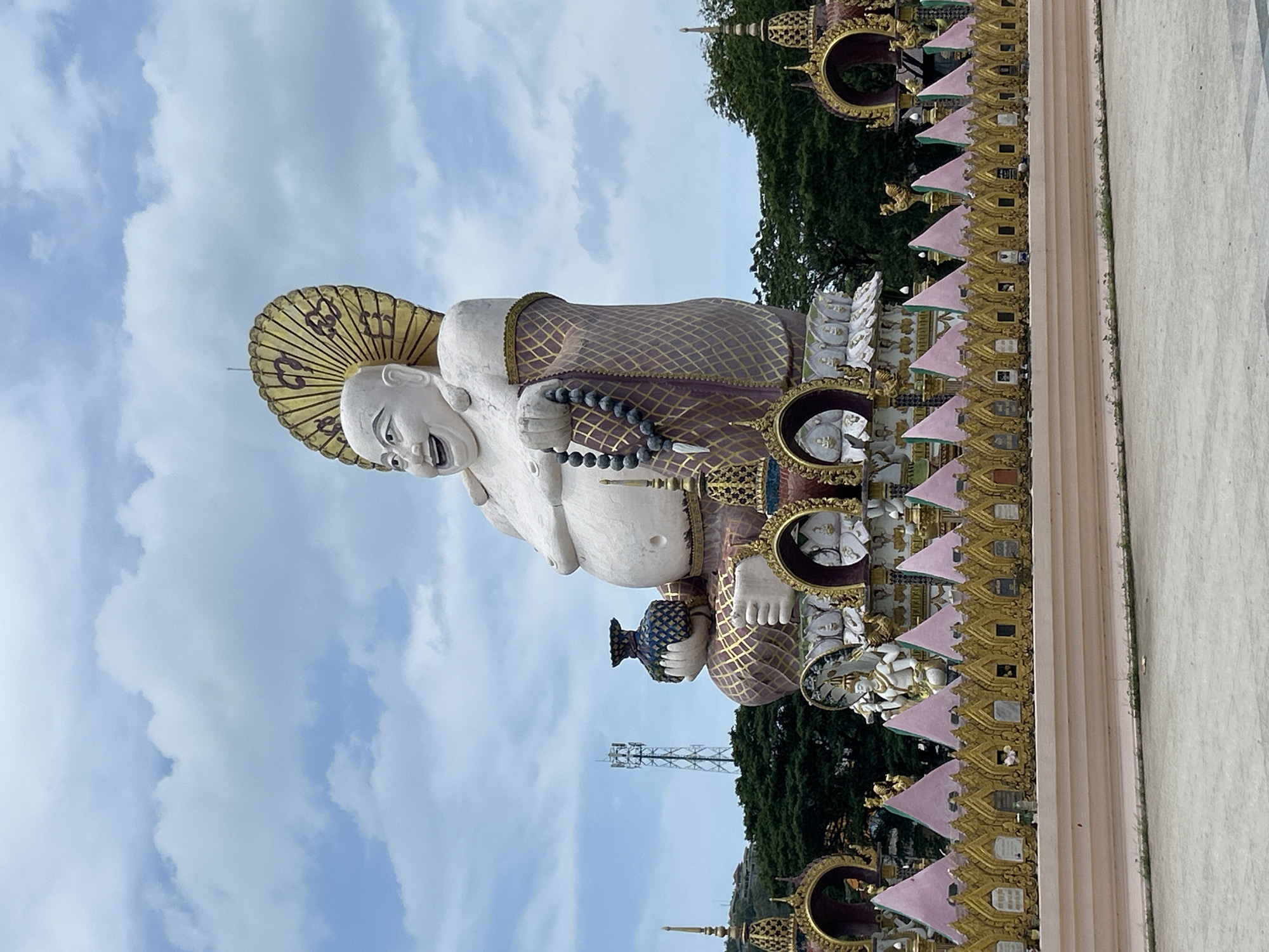 A monumental, white, seated Buddha statue with a large, jovial expression and a golden umbrella-like halo stands against a cloudy blue sky. The Buddha is adorned with a patterned robe and a string of large beads. In front of the Buddha is a decorative golden fence with pink triangular accents and small arched niches. Behind the fence, other smaller statues and decorative elements are visible, including a multi-armed statue. Trees and a cell tower are in the background. The scene is outdoors, likely in a public space or temple complex on Ko Samui District, Thailand.