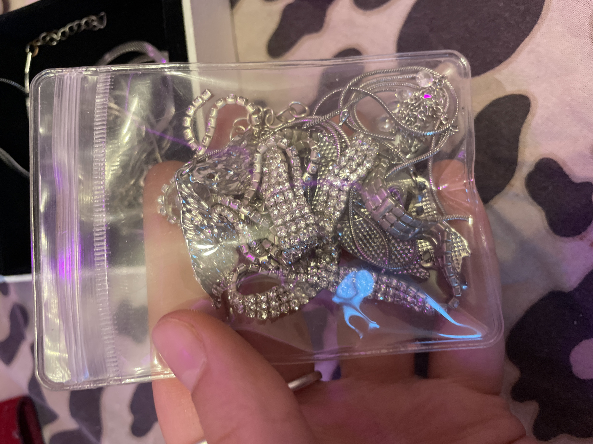 The image shows a close-up of a hand holding a clear plastic bag filled with silver-colored jewelry. The jewelry appears to be a tangle of necklaces, bracelets, and possibly charms, many of which are adorned with small, sparkling stones that reflect the purple and blue lighting in the scene. The background is blurred but shows a patterned surface, possibly fabric or wallpaper. There is no visible indication of the city of Ladozhskoe Ozero, Russia, nor any other specific location. The image is a static shot, focusing on the contents of the plastic bag.