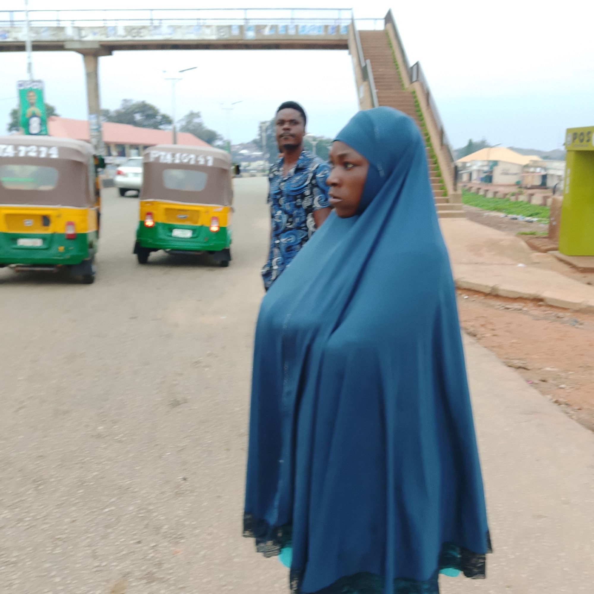 In a city street, three-wheeled vehicles and a pedestrian bridge are visible. The image features a woman wearing a blue hijab, a man in a patterned shirt, and two auto-rickshaws. One auto-rickshaw, yellow and green, has the license plate "TA-7274". The second auto-rickshaw, also yellow and green, displays "PTA10157". In the background, buildings and a pedestrian walkway with stairs are present. The ground is paved with asphalt.
