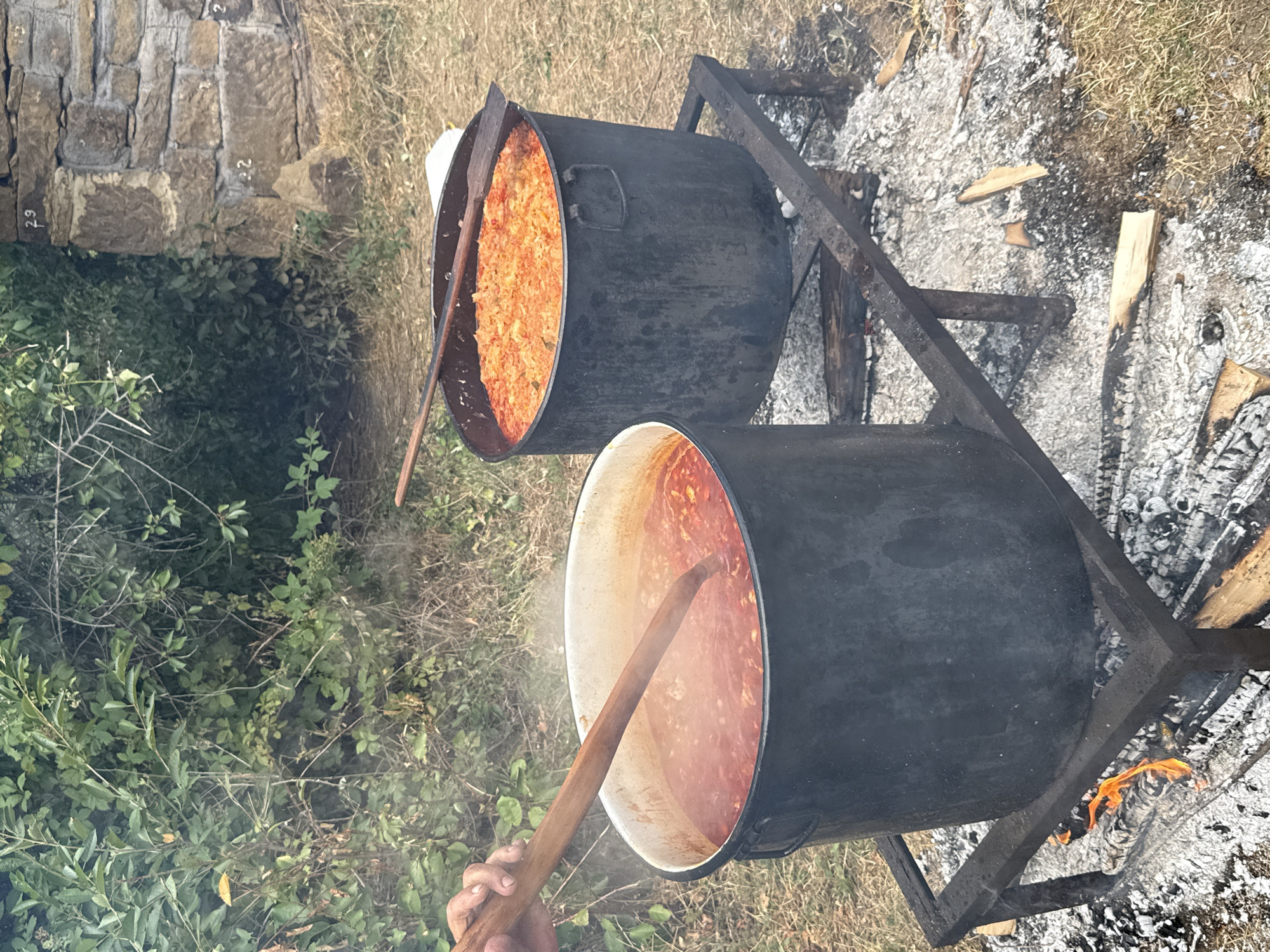 This image depicts an outdoor cooking scene, likely in Manaselska Reka, Bulgaria. Two large, dark pots are placed on a metal grate over a fire, indicating a rustic or communal cooking setup. The pot in the foreground, with a white rim, contains a simmering red stew, and a wooden spoon is being used to stir it. A hand is visible holding the spoon. To the right, another dark pot holds a dish with orange-colored ingredients, possibly cabbage or a similar vegetable. The background features dense green foliage and a section of a stone wall with numbered markings ("29" and "22"), suggesting it might be part of an outdoor structure or historical site. The ground is covered in dry grass and ash from the fire.