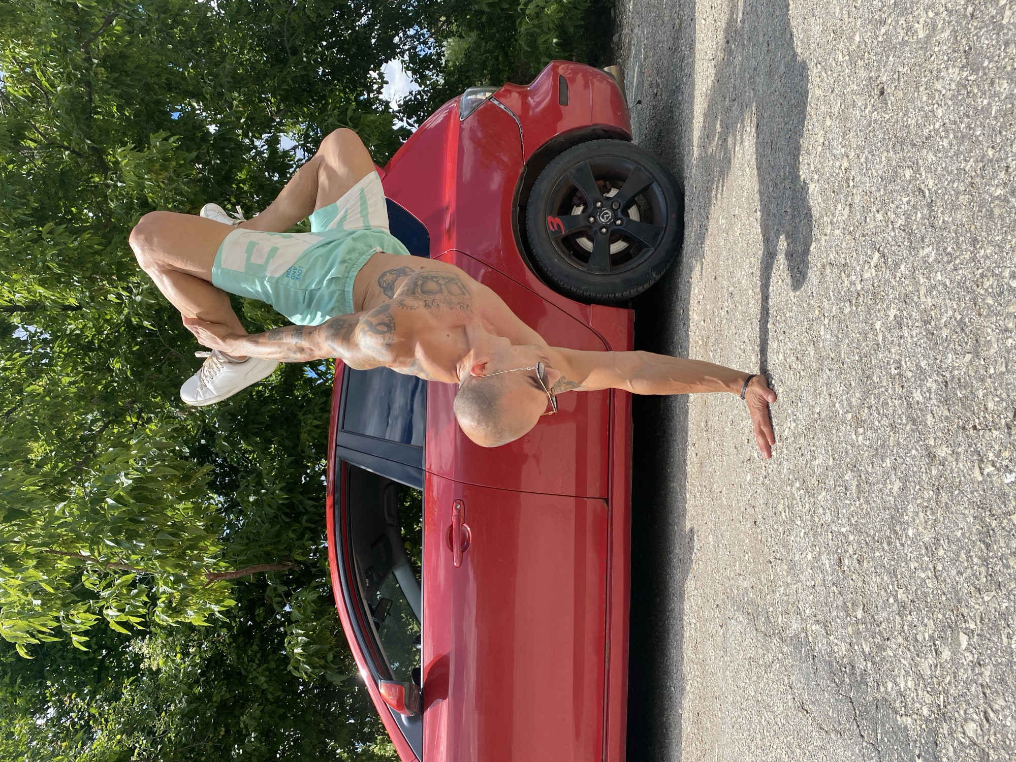 A shirtless, muscular man with tattoos is performing a handstand next to a red car in Pleven, Bulgaria. He is wearing light green shorts and white sneakers. The man's body is inverted, with his legs bent and crossed in the air. His right hand is supporting his weight on the ground, while his left arm is bent behind his back. The red car is parked on a gravel surface, with trees and foliage in the background. The sky is clear and blue, suggesting it is a sunny day.