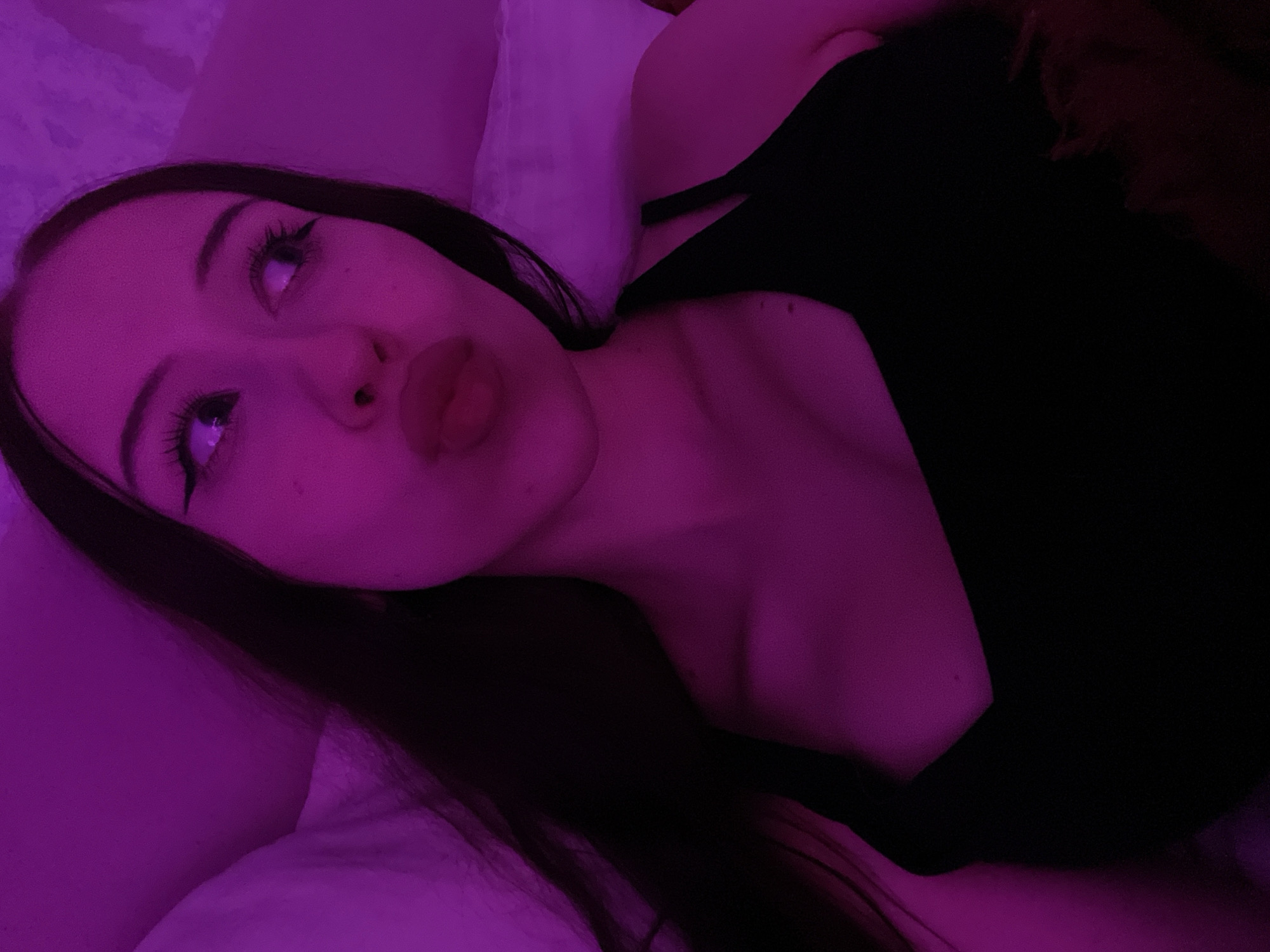 The image shows a young woman with dark hair wearing a black top. She is lying down, possibly in bed, with a pillow behind her. The lighting in the image is predominantly purple, creating a moody and atmospheric effect. Her facial expression is one of playful pouting, with her lips pursed. Her eyes are looking upwards and to the side, and she has dramatic winged eyeliner. The overall impression is of a selfie taken in a bedroom environment. There is no information in the image to suggest a location in Votkinsk, Russia.