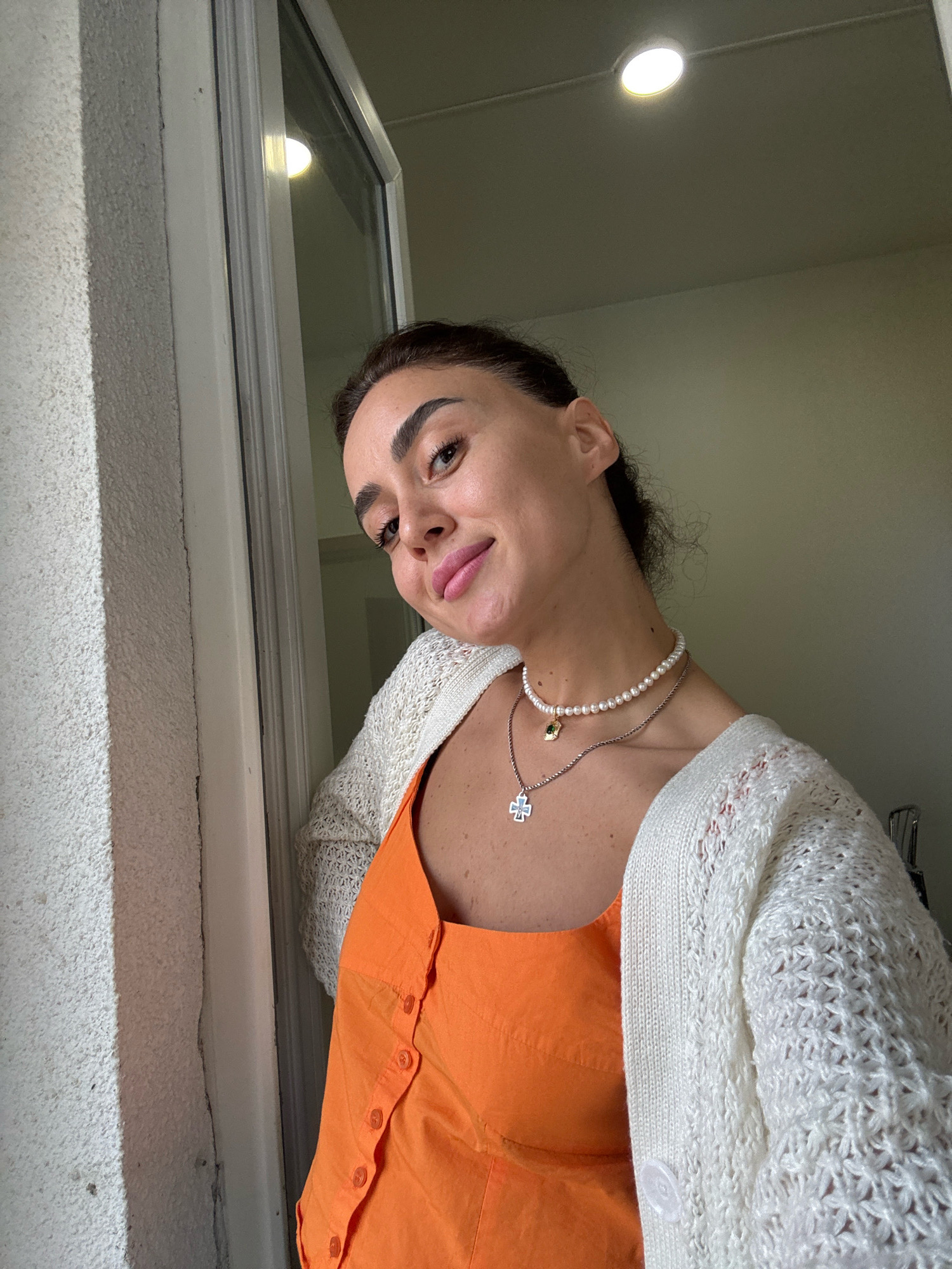 A woman with dark hair pulled back is pictured from the chest up, looking toward the viewer with a slight smile. She is wearing an orange dress and a white knitted cardigan. She has two necklaces on, one with pearls and a small green pendant, and another with a cross pendant. She is standing next to a window. The scene appears to be indoors, with natural light coming from the window and overhead lighting visible. The exterior wall of a building is visible to the left, suggesting an urban setting, potentially in Tbilisi, Georgia given the context.