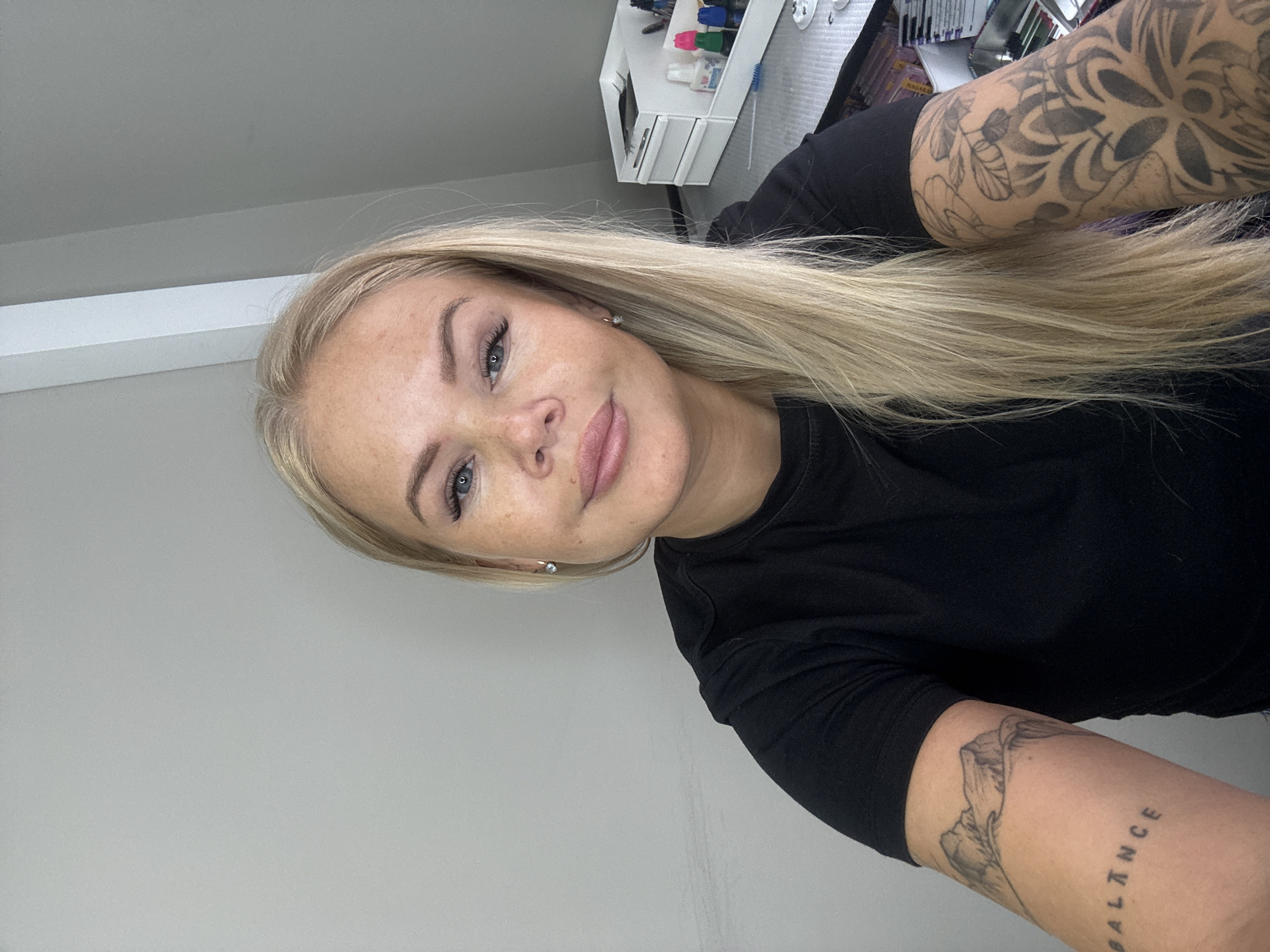 A woman with blonde hair, wearing a black long-sleeved shirt, is facing the camera. She has fair skin, blue eyes, and freckles. Her left arm, closer to the camera, has a tattoo that includes the word "BALANCE". Her other arm, partially visible in the upper right corner of the frame, has a floral tattoo. The background is a light gray wall on the left, and a desk with various items on the right. The location is Unknown, Unknown.