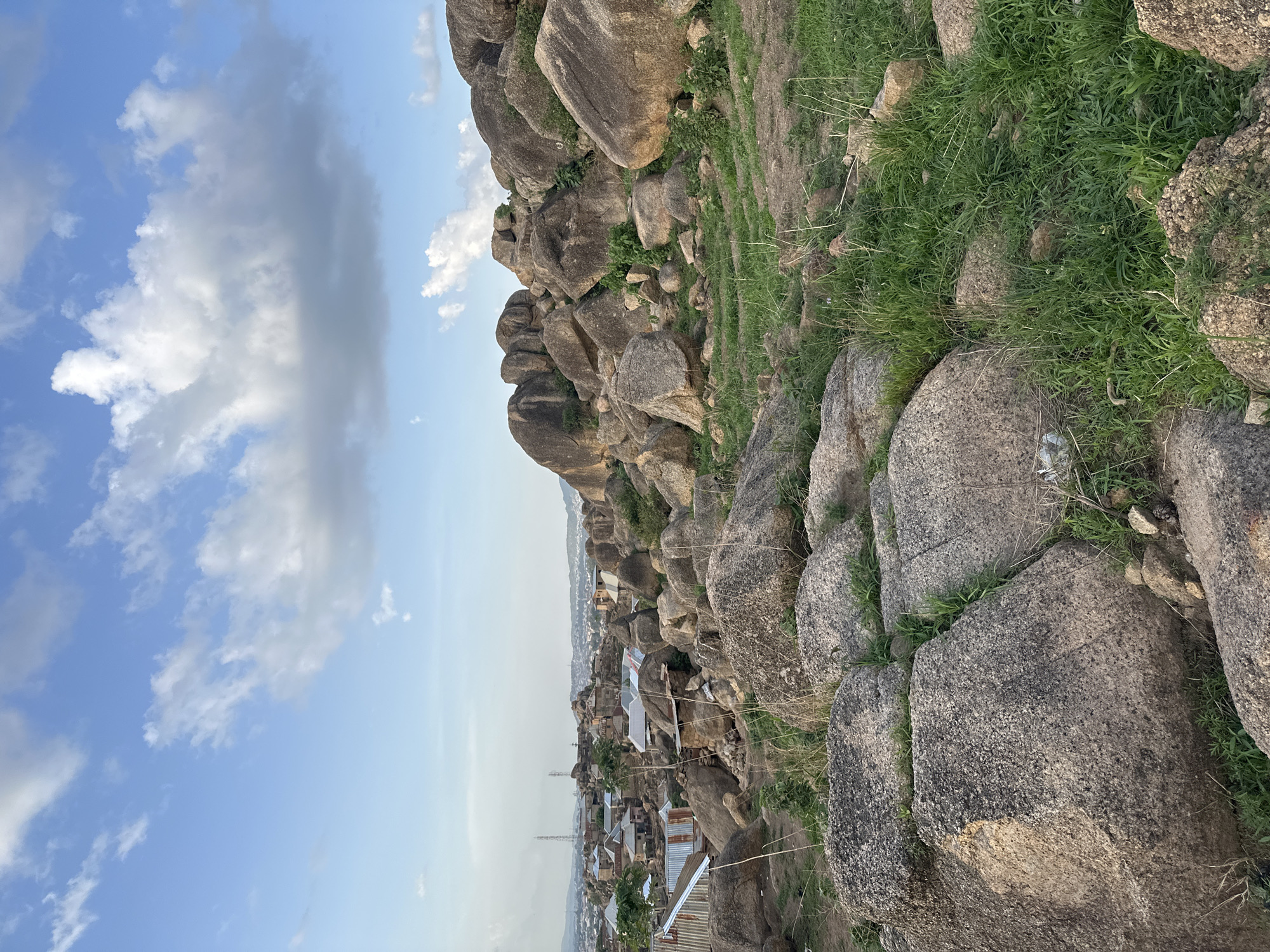 The image captures a panoramic landscape view from a rocky hillside overlooking a town in Jos, Nigeria. The scene is dominated by natural elements, with large, irregularly shaped boulders of grey and brown hues scattered across the foreground and mid-ground. Patches of green grass and some drier vegetation grow in between and around these rocks. Closer inspection reveals a small amount of litter, such as a crumpled plastic item, caught among the rocks and grass in the lower right section.
The rocky terrain slopes down towards a sprawling settlement. The town below consists of numerous buildings, predominantly residential, characterized by a mix of silver and rusty corrugated iron roofs. These structures appear densely packed, creating an urban or semi-urban environment. A tall, slender lattice tower, possibly for telecommunications or electricity, is visible rising above the rooftops in the mid-distance within the town.
The background features a wide expanse of clear blue sky, adorned with scattered white and light grey cumulus clouds, suggesting fair weather. The horizon line above the distant town appears slightly hazy. The lighting indicates it is daytime, with soft, natural illumination casting gentle light across the landscape. There are no people visible in the image, nor any specific activities or events taking place. No visible text is present.
