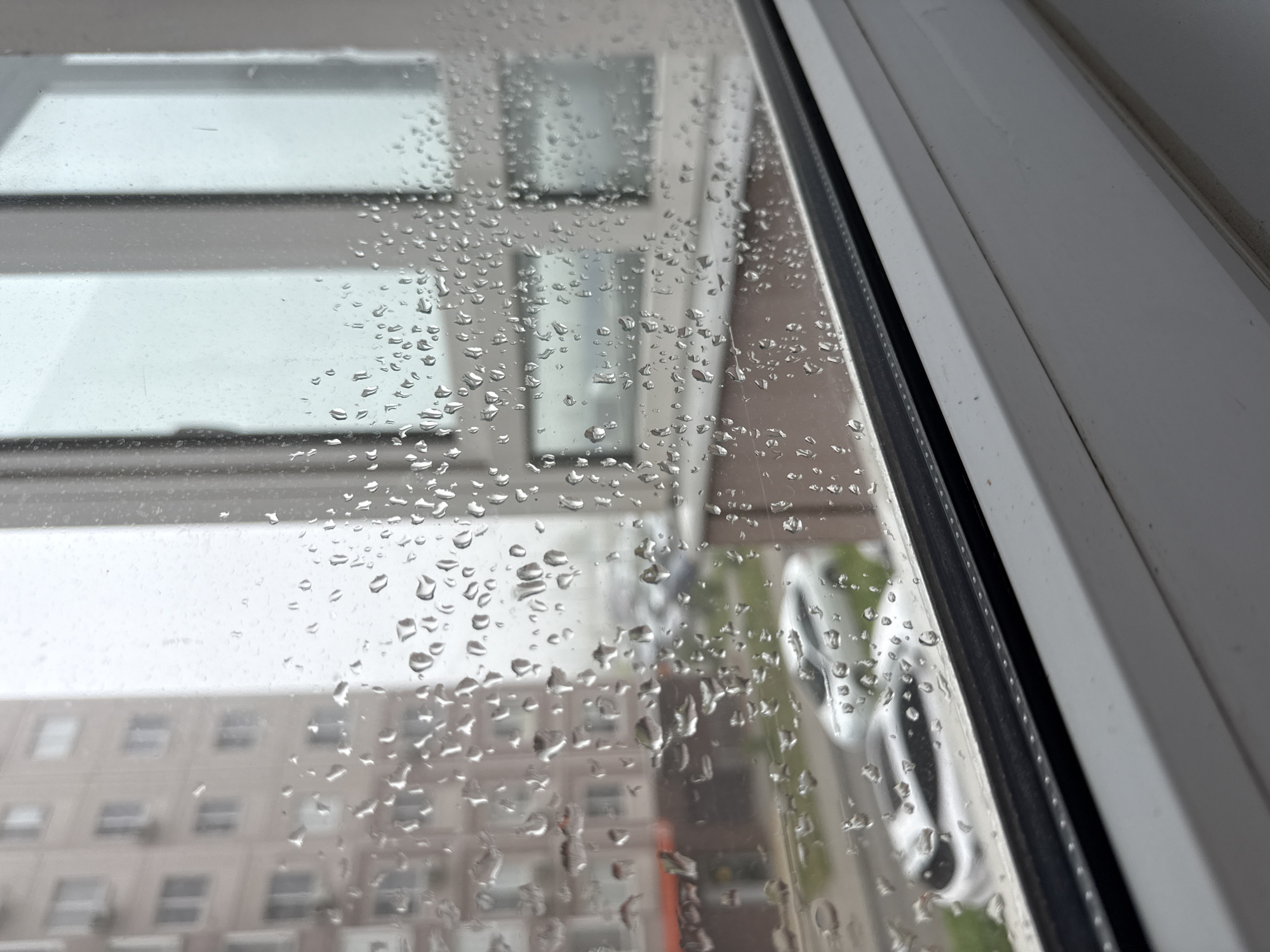 The image shows a view through a rain-covered window. Outside, a multi-story building is visible, suggesting an urban environment. The building appears to have many windows, indicating a residential or office structure. A vehicle is also partially visible through the raindrops, further supporting the urban setting. The window frame is white with a black weather stripping. It is raining. The scene is probably in Rostov-on-Don, Russia.