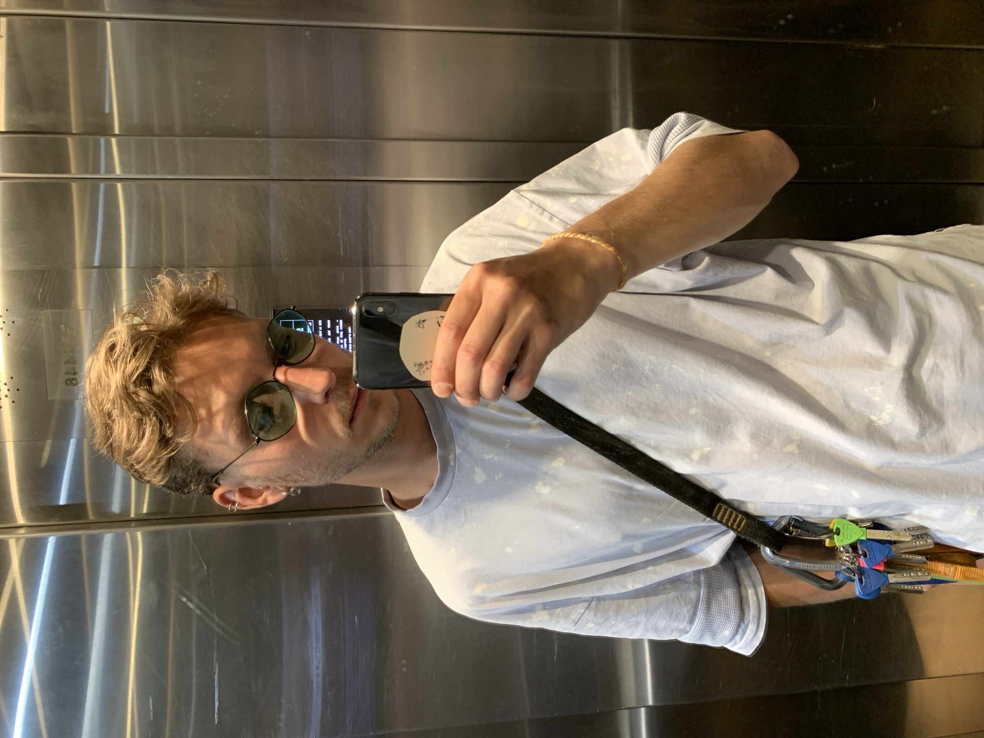 A male subject takes a selfie in a metallic elevator. He is fair-skinned with brown hair, wearing sunglasses and a light blue t-shirt. A black shoulder strap with climbing equipment attached is visible across his body. The interior of the elevator is stainless steel. The location is unknown, and there's no evidence suggesting this scene is in Tel Aviv, Israel.