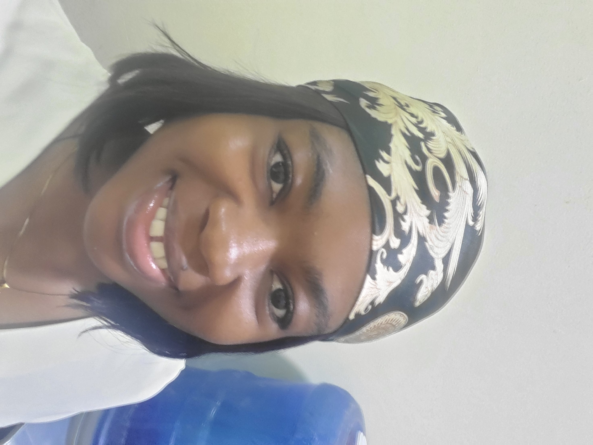 A young woman is photographed indoors. She has dark skin and short black hair with a patterned headscarf. She is smiling with a clear view of her teeth. Part of a blue water cooler is visible in the foreground. The location is Gwagwada, Nigeria.