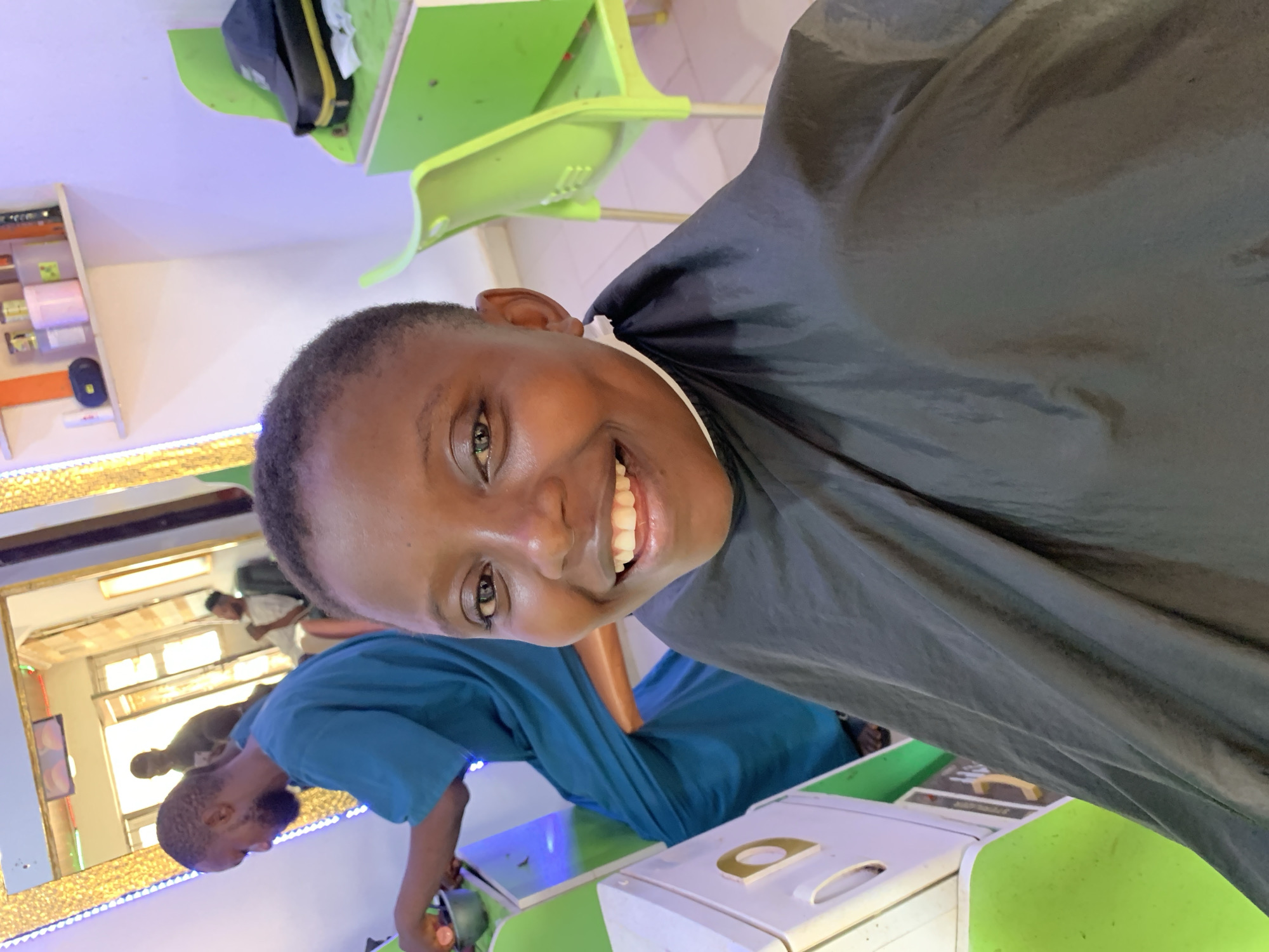 A young person with short, dark hair is smiling at the camera while wearing a black cape, likely at a barber shop in Ikorodu, Nigeria. The person is seated, with a table in front of them covered in green paper. On the table is a white device with a gold design. A barber is visible in the background, wearing a blue shirt and working on another person reflected in a mirror with a decorative gold frame, indicating a professional setting. In the top left, there are hair products on shelves and a black bag on a green shelf. The overall environment appears to be a brightly lit, clean, and functional business establishment.