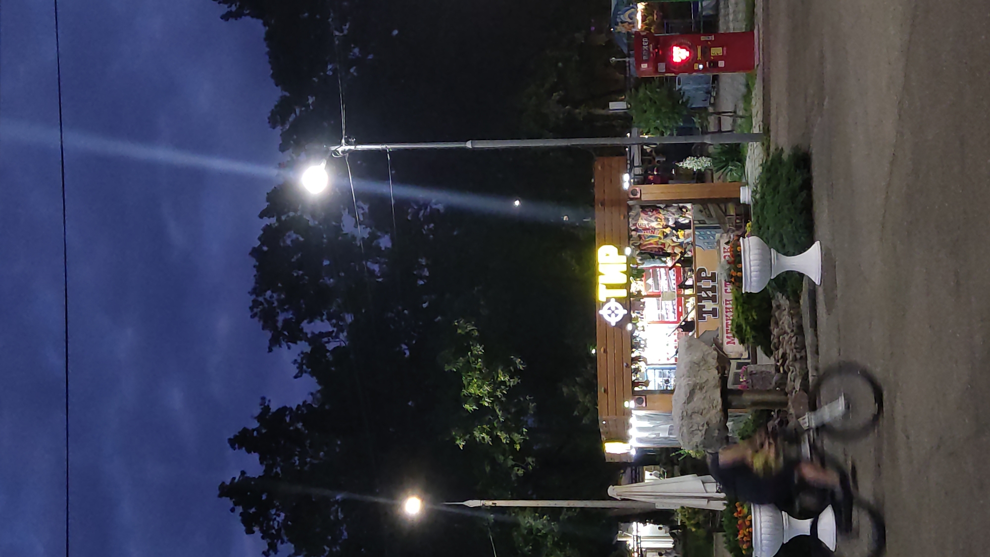 In the city of Krasnodar, Russia, a street scene unfolds at dusk. The sky is a deep, cloudy blue, with a single streetlamp casting a bright beam downwards. In the foreground, a person on a bicycle is captured in motion blur, moving from left to right across the frame. To the right of the cyclist, a small wooden kiosk with a neon sign reading "ТИР" (which translates to "Shooting Gallery") is illuminated. Various items, possibly prizes, are visible inside the kiosk. Further to the right, a red carnival-style game, likely a strength tester, is also lit up. The scene is lined with trees and two decorative white planters filled with greenery and flowers. The overall impression is of a lively park or entertainment area as evening sets in.