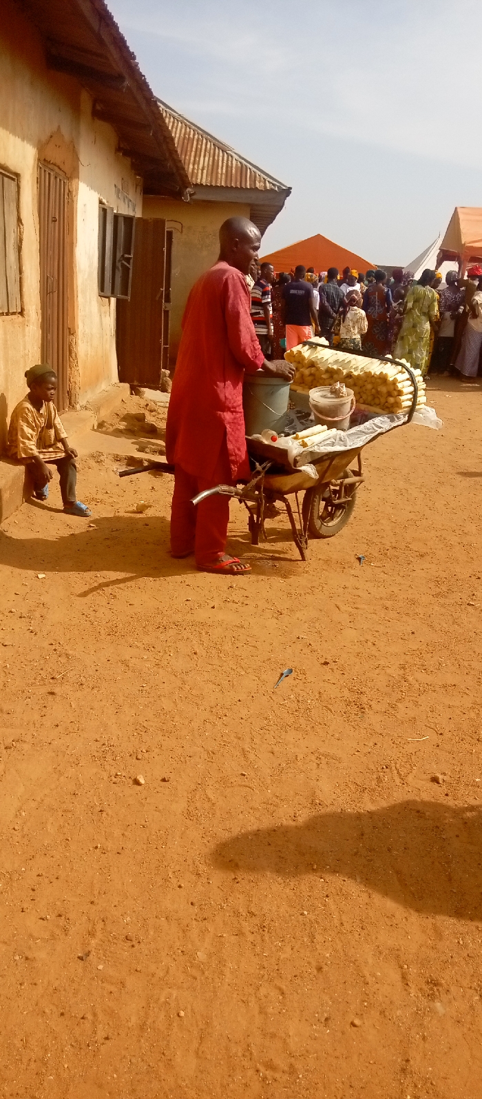 The image captures an outdoor scene in Kasuwan Magani, Nigeria, under a clear, hazy sky. The ground consists of reddish-brown dirt, with a metallic spoon-like object and a small blue item visible on its surface. In the center, a man in a red tunic and trousers, wearing red sandals, stands with his back partially to the viewer, facing a two-wheeled pushcart. The cart is laden with goods, including a grey bucket, a lidded plastic container, a white plastic bottle, and multiple rows of horizontally stacked, cylindrical, pale yellow wrapped food items. To the left, a young person dressed in a light brown shirt, dark trousers, and light blue sandals sits on a raised, light-colored step of an adjacent building. This building features a weathered wooden door and open windows with wooden grilles, topped by a corrugated metal roof. In the background, a large group of people is gathered, and several tents, including a prominent orange canopy, are visible above the crowd.