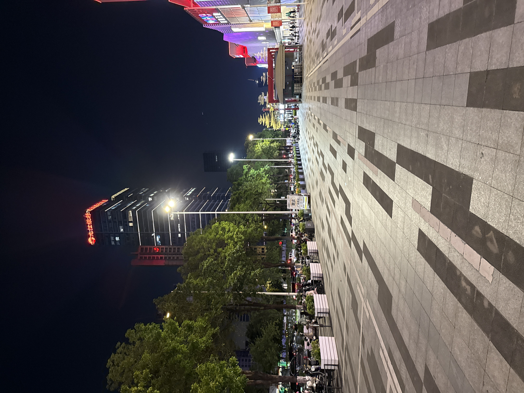 This image captures a street scene at night in Tianhe District, China. A tall skyscraper with illuminated windows and a red sign reading "九江银行" (Jiujiang Bank) dominates the background. The street is paved with rectangular tiles arranged in a geometric pattern of light and dark shades.
On the left side of the street, a line of trees with lush green foliage is visible, separating the pedestrian walkway from a row of parked vehicles, including cars and many scooters. Planters with green plants are placed along the sidewalk. Several people are seen on the left side, some near the parked vehicles, and others walking.
The right side of the street features modern buildings with colorful illuminated facades, suggesting commercial establishments or shopping centers. A small, covered structure resembling a kiosk or information booth is situated in the middle ground. More people are walking on the right side of the street. Streetlights illuminate the scene, casting light on the pavement and surrounding areas. The overall atmosphere is one of urban activity at night.