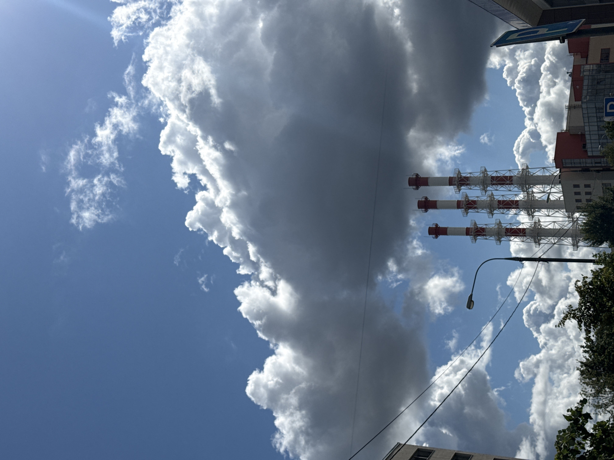 The image depicts a clear, sunny day in Lyubertsy, Russia, with a bright blue sky partially obscured by large, fluffy white and dark gray clouds. In the foreground, several industrial smokestacks with red and white banding stand tall, emitting plumes that are not visible. A street lamp is positioned in front of these structures. The lower part of the image shows the roofs and upper floors of several buildings, some of which appear to be industrial or commercial, with red and white accents. Dense green foliage from trees can be seen at the bottom of the frame, and overhead power lines crisscross the sky. A blue sign with a white "P" on it, indicating parking, is visible on the right side of the image. There are no people visible in the scene.