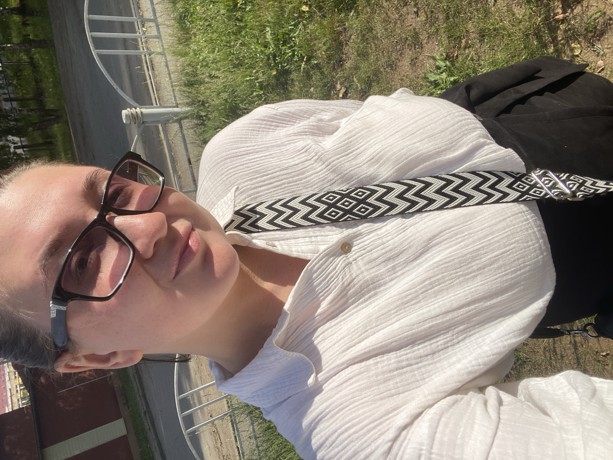 A woman with dark hair pulled back poses for a selfie outdoors. She wears glasses and a white, crinkled button-up shirt. A black bag with a patterned strap hangs across her chest. She is wearing a black skirt. The background shows a white picket fence, grass, trees, and a brick building. The sunlight casts shadows on her face and shirt.
