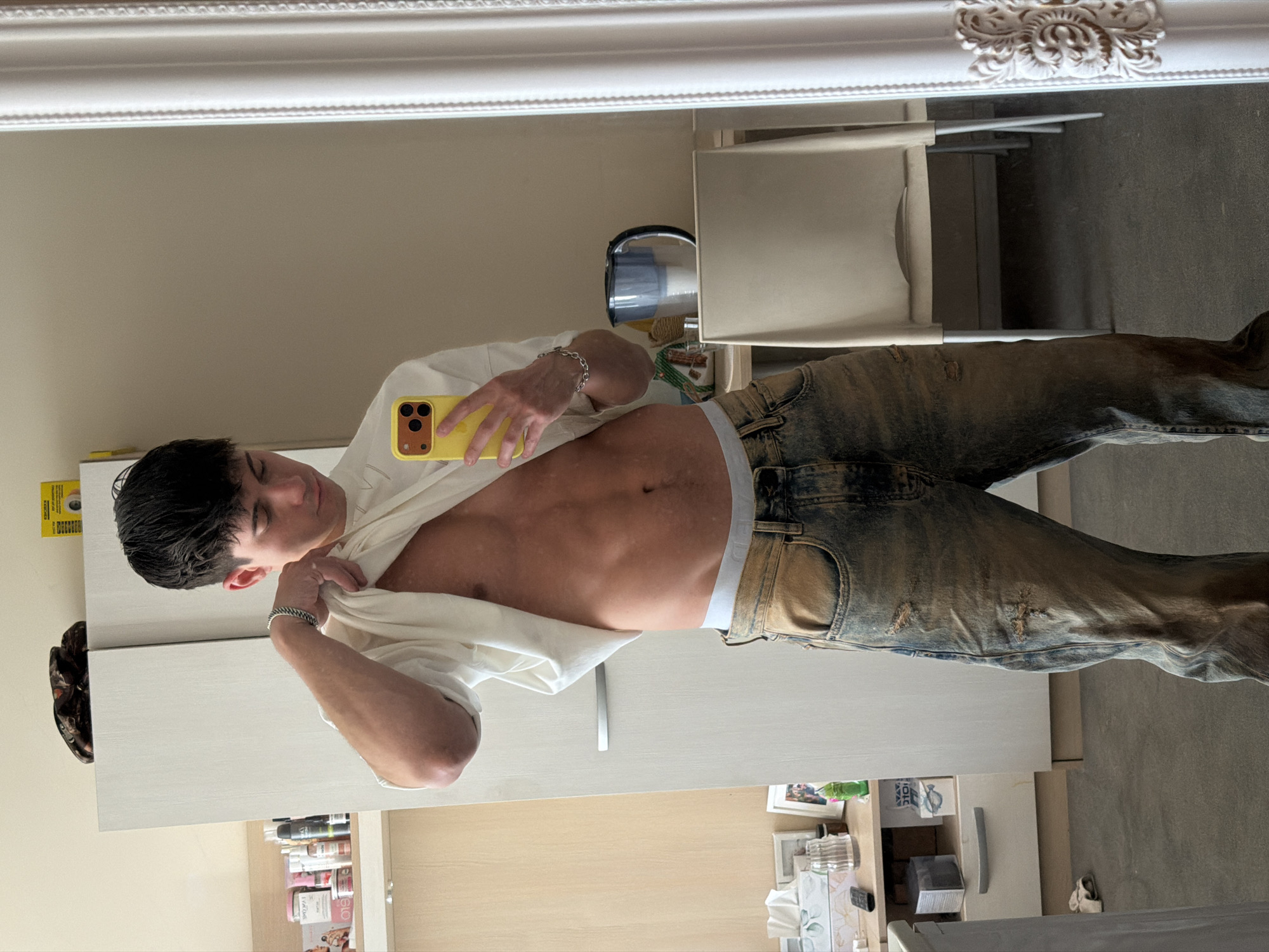 A young man is taking a selfie in a mirror, holding a yellow phone in his right hand. He is wearing distressed jeans and a white t-shirt that he has partially lifted to expose his abdomen. The reflection shows him in what appears to be a bathroom or bedroom with a mirror, vanity, cabinets and drawers. The surrounding objects include toiletries and other personal items. There's no indication of a location in the city of Sofia, Bulgaria.
