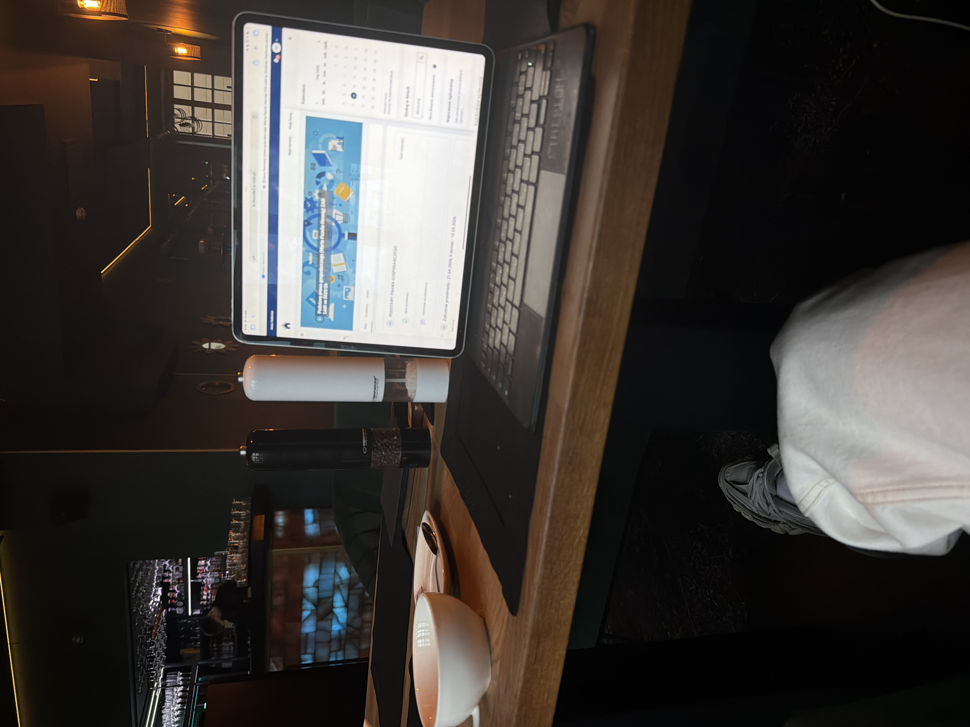 A close-up shot shows a tablet with a keyboard attachment on a wooden surface, possibly a table or desk. The tablet displays a screen with what appears to be a calendar or task management application. To the left of the tablet, there's a white cylindrical object and a black, cylindrical object. A cup or mug rests on a saucer near the bottom of the frame. The background is dark, with hints of a dimly lit interior.
On the right edge of the frame, a person is partially visible, wearing white clothing and grey sneakers. The shot appears to be taken in an indoor setting, maybe a cafe or bar, as suggested by the bar glasses in the background.