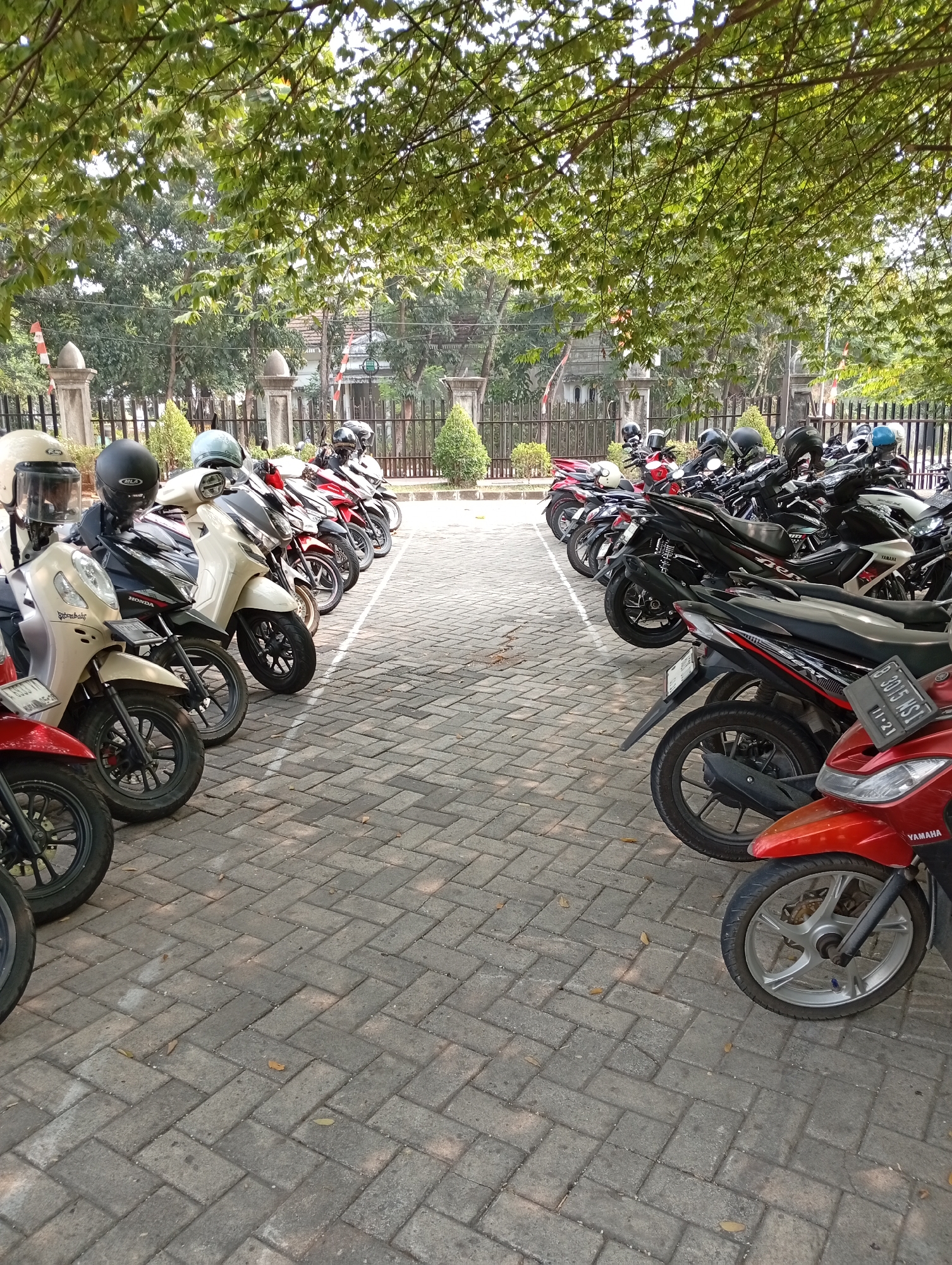 The image depicts a row of parked motorcycles under the shade of trees, on a paved area made of grey interlocking bricks. The motorcycles are predominantly dark-colored with some red and white accents. Most of them are scooters, and some have helmets placed on their seats or handlebars. The environment appears to be an outdoor parking lot in a semi-urban or suburban area, with a fence and some buildings visible in the background. The lighting suggests it is daytime, with dappled sunlight filtering through the leaves. The scene is static, with no people actively interacting with the motorcycles. This location is identified as being in Kelapa Dua, Indonesia.