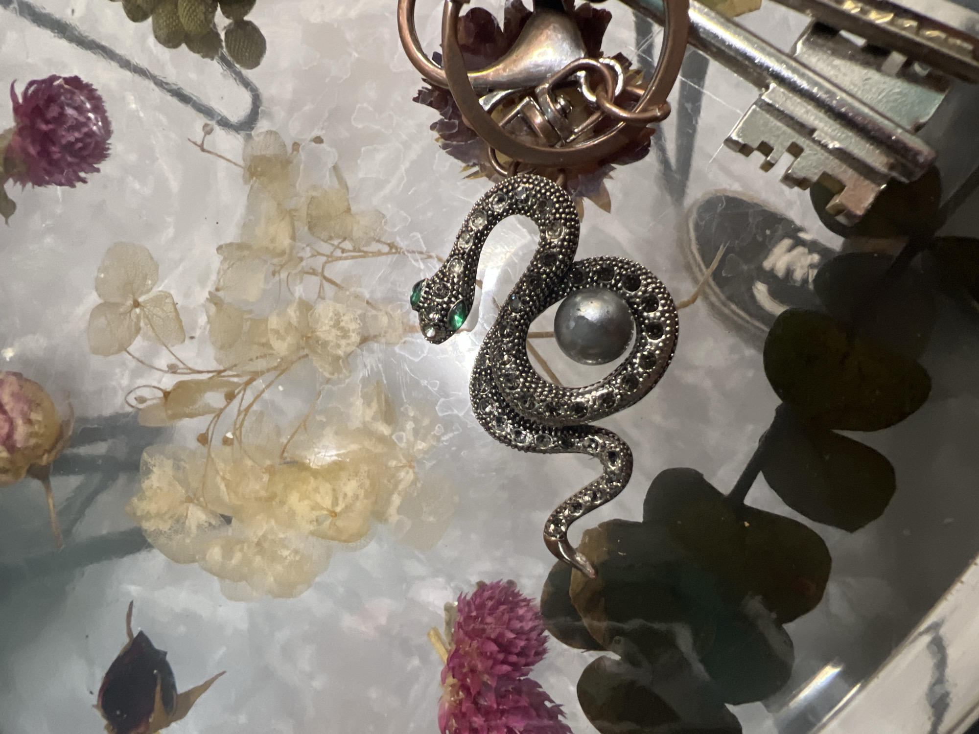 A close-up, top-down shot showcases a collection of dried flowers, a snake figurine adorned with small clear and green stones, and a large grey pearl. The snake is coiled, with its head and tail raised, and its body is encrusted with tiny reflective stones. The pearl is positioned near the center of the snake's coil. In the background, various dried floral elements are visible, including pale yellow and pinkish-red blooms, as well as dark green leaves. To the right, a copper-colored object with a circular design and part of a key are also visible. The scene appears to be an indoor still life arrangement, possibly on a glass surface given the reflections. There are no people or any indications of activity or interaction. There is no information available to determine the location in the city of Ленинский, Russia.