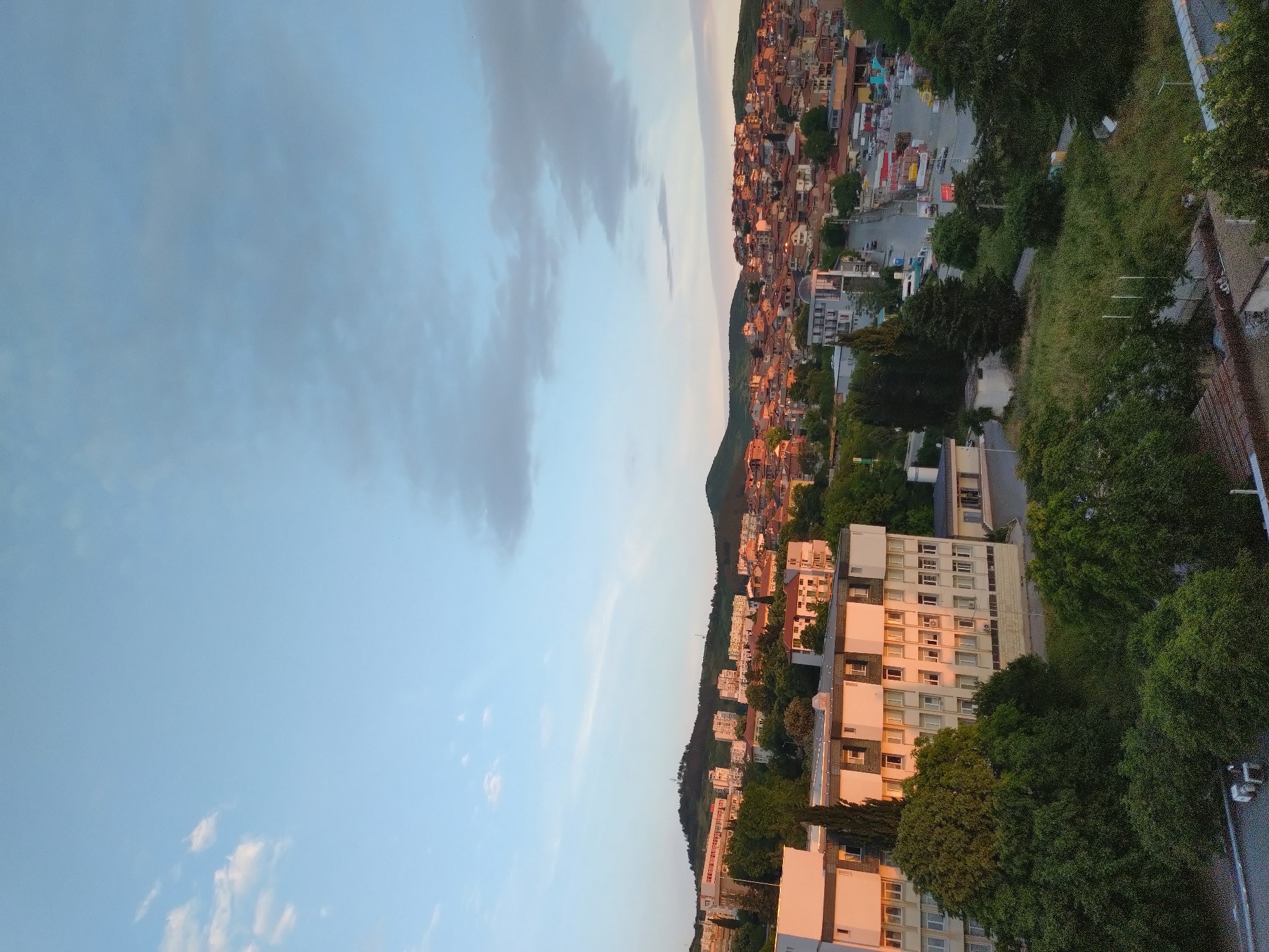 The image shows a landscape view of Stara Zagora, Bulgaria, during sunset or sunrise. The scene is divided between the sky on the left and the city and nature on the right. The sky is filled with light blue and gray clouds. The city buildings cluster on a hillside and along the lower slopes. Green trees are interspersed with the buildings, especially along the lower right edge of the image. A series of multistory apartment buildings are visible in the lower portion of the image. The sunlight casts a warm glow on the buildings and the sky.