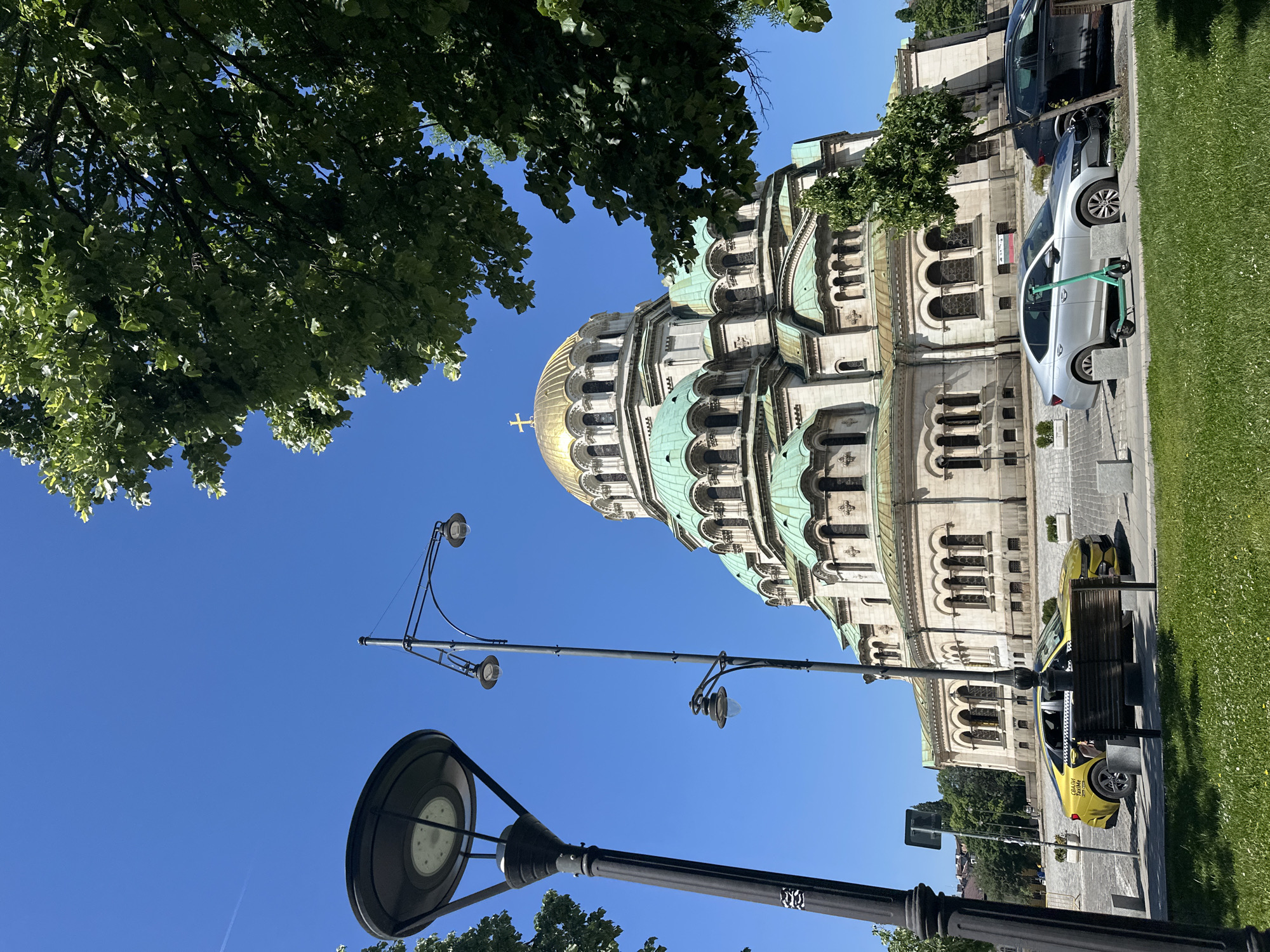 This image shows the Alexander Nevsky Cathedral in Sofia, Bulgaria. The cathedral is an elaborate structure with a gold-plated dome and several smaller green domes. The sky is a clear blue. In the foreground, there are lampposts and tree branches. Parked cars and a section of green lawn are visible.
