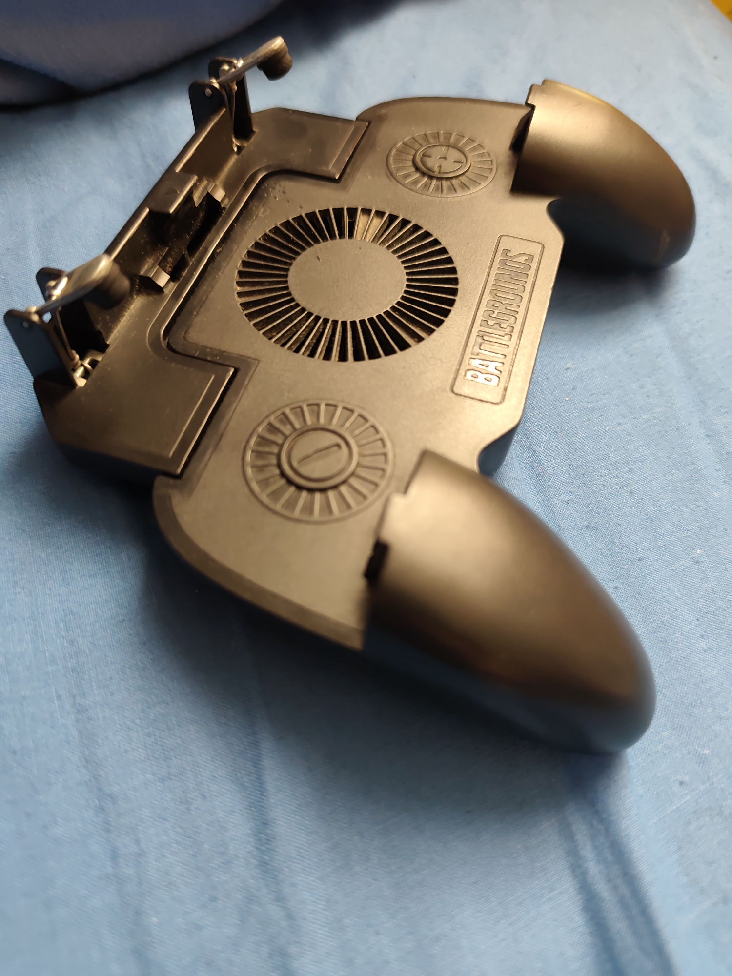 A black, plastic mobile gaming controller with a built-in fan is positioned on a light blue textured fabric surface. The controller has grips on both sides, a fan vent in the center, and two trigger-like buttons at the top. The text "BATTLEGROUNDS" is embossed on the surface of the controller. The lighting suggests it is indoors, with shadows cast by the controller onto the fabric. There are no people or interactions visible. The image does not provide any information about the location in the city of Stare, Ukraine.