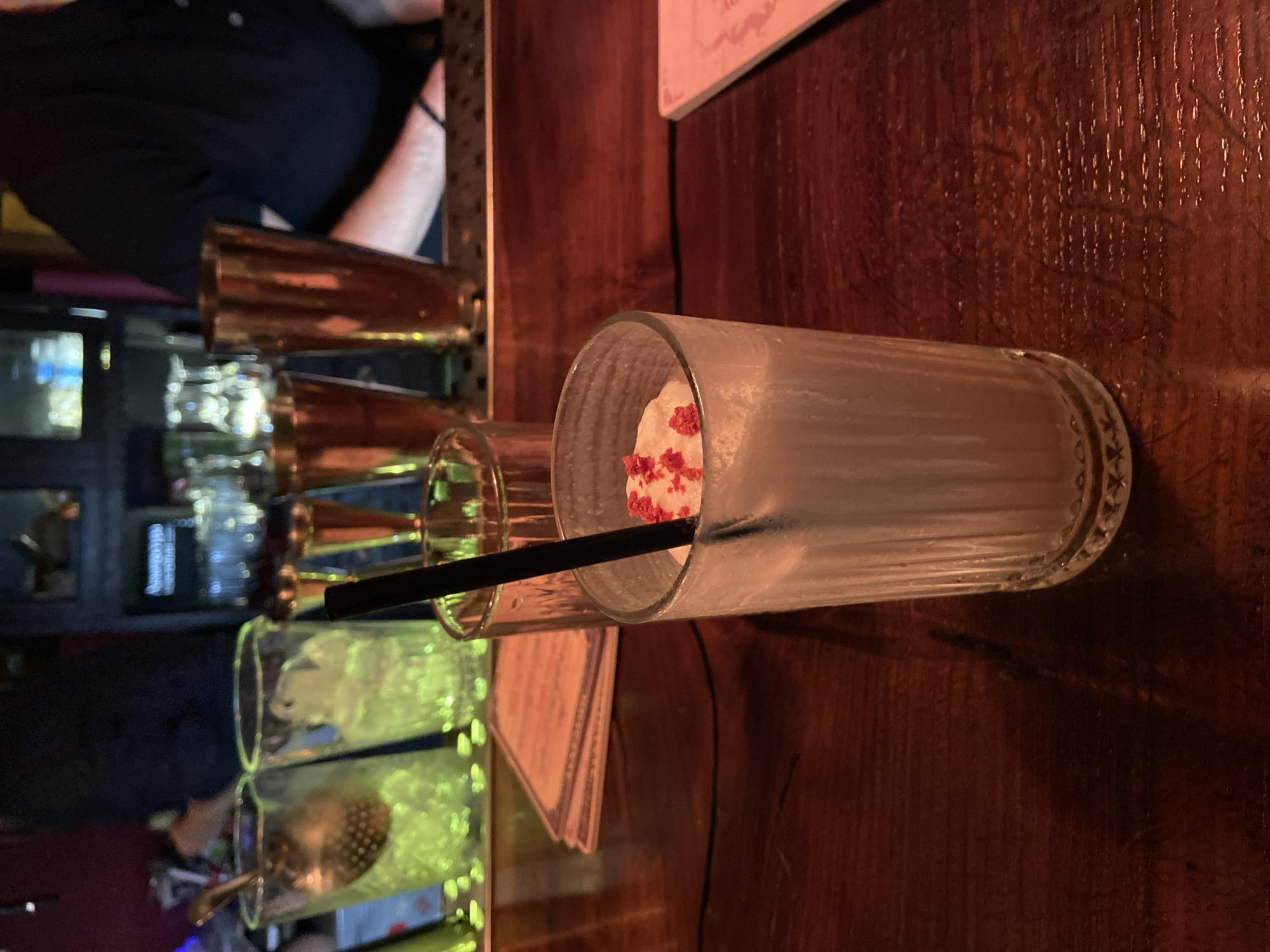 A drink sits in a ribbed glass in the foreground, topped with whipped cream and red sprinkles and a black straw. The drink is on a dark wooden bar. In the background, there are several glasses, some filled with ice, and metal cocktail shakers. A person wearing a black shirt is visible behind the bar. The image does not contain any information that would allow for the location in the city of Brest, Belarus to be identified.
