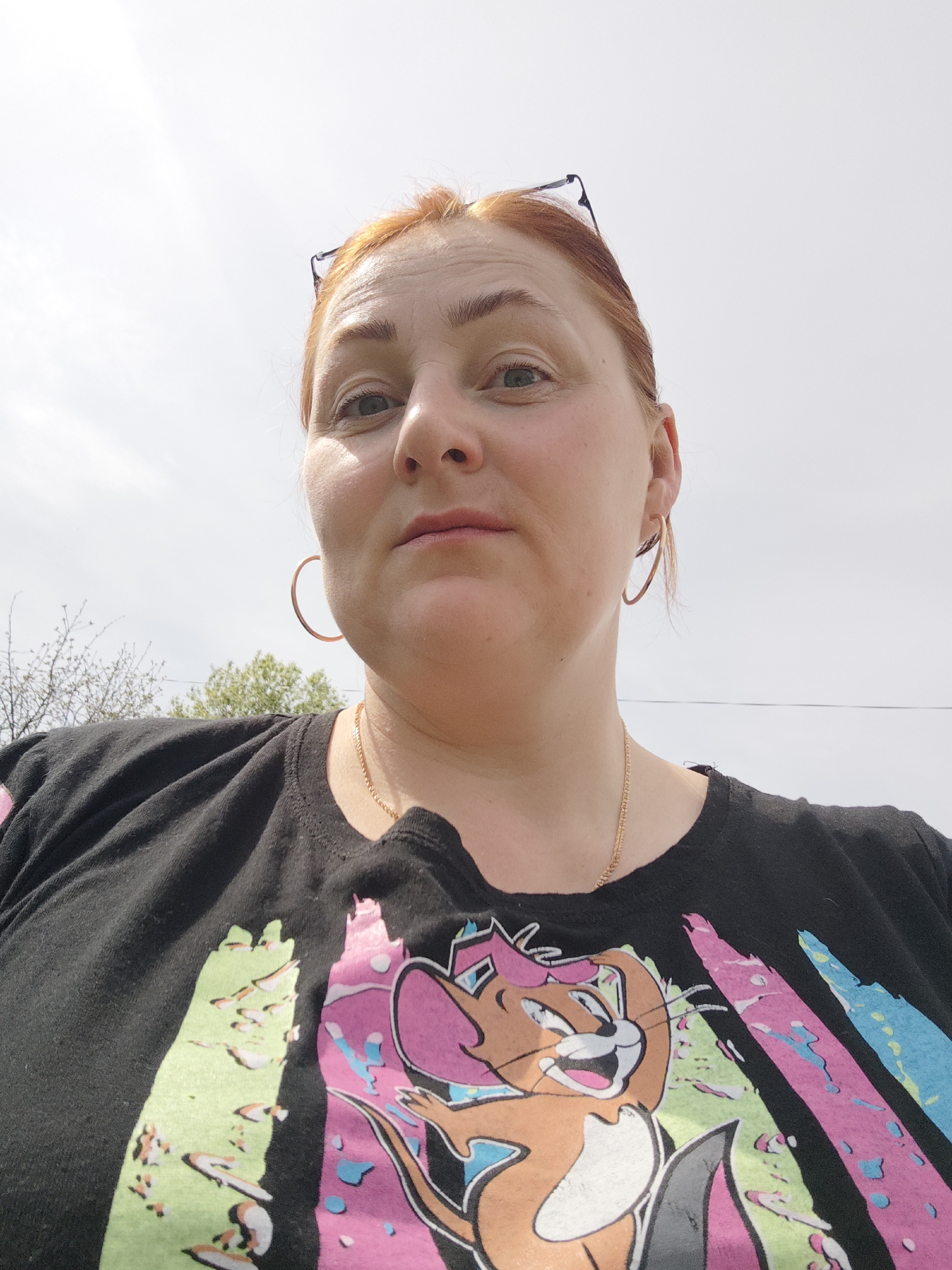 A woman with fair skin and red hair is facing the camera. Her glasses are perched on her head, and she is wearing gold hoop earrings and a gold necklace. She is wearing a black t-shirt with a colorful graphic design. The background is an overcast sky and the branches of trees are visible in the distance. The image was taken in the city of Большая Уса, Russia.