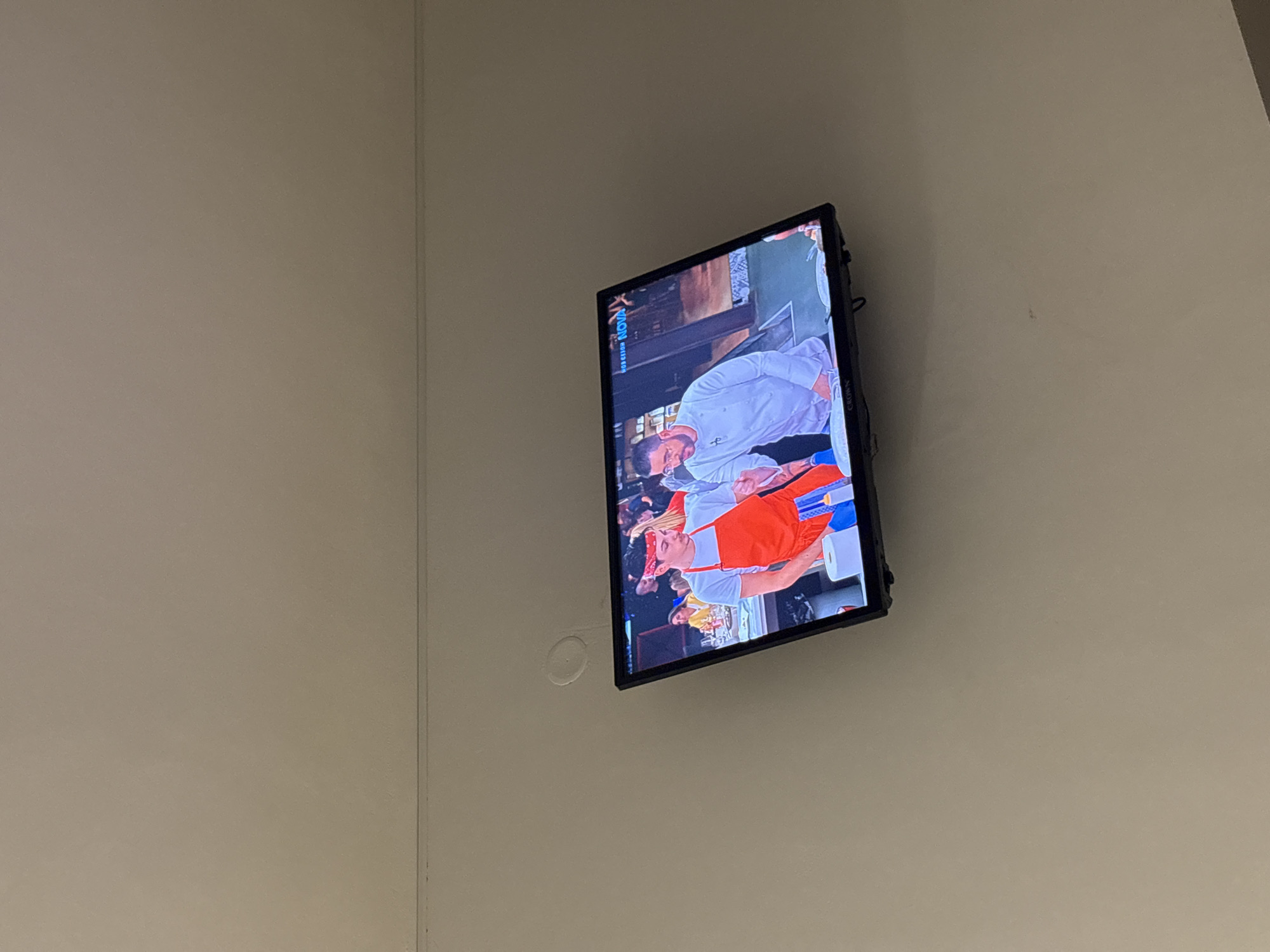The image shows a wall-mounted television displaying a scene with two people. One person wears a white shirt and the other is wearing an orange apron. The location is indoors, in a room with light-colored walls. The image does not include details that confirm a specific location in Sofia, Bulgaria.